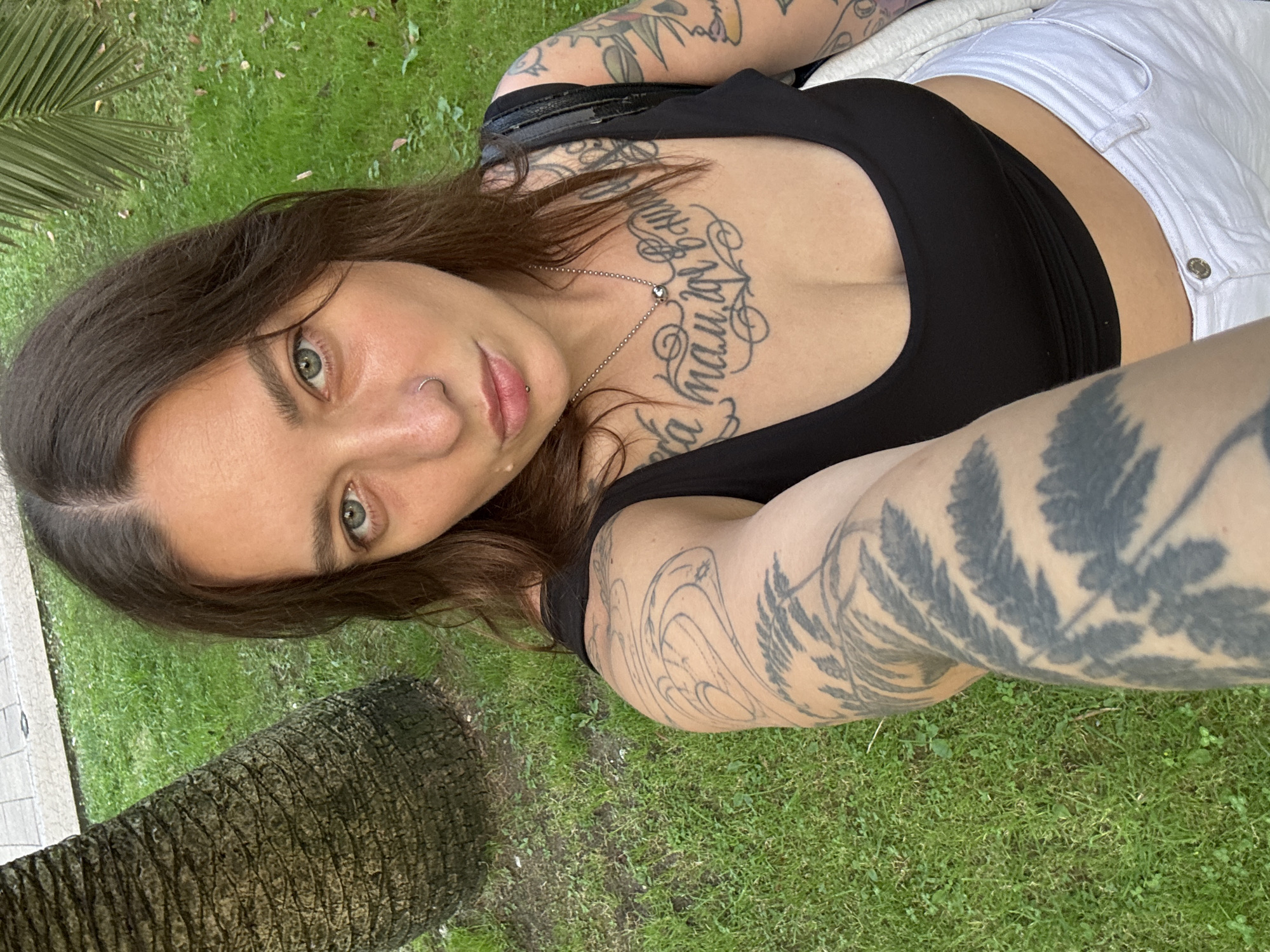 A woman with brown hair, blue-green eyes, and a nose ring takes a selfie outdoors. She is wearing a black crop top and white jeans. The background shows a palm tree trunk, green grass, and a palm frond. The woman has tattoos on her arms and chest.