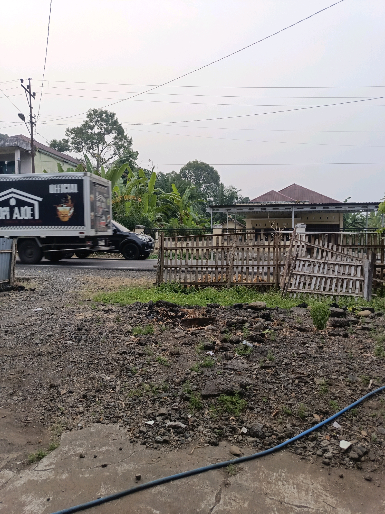 The image depicts a street scene in Ulak Tanding, Indonesia. In the foreground, a rough, gravelly ground transitions to a concrete path where a blue hose lies. Behind this, a low, weathered wooden fence borders an area with sparse vegetation and debris. Further back, a dark-colored truck with a black box body, featuring a white logo and text that appears to read "KOPI AJOE OFFICIAL," is in motion on a paved road. To the right of the road, a house with a red tile roof and a covered porch is visible, with a wooden fence in front of it. Lush green banana trees and other foliage are interspersed throughout the scene, and utility poles with electrical wires stretch across the overcast sky. The overall impression is of a rural or suburban street in Indonesia.