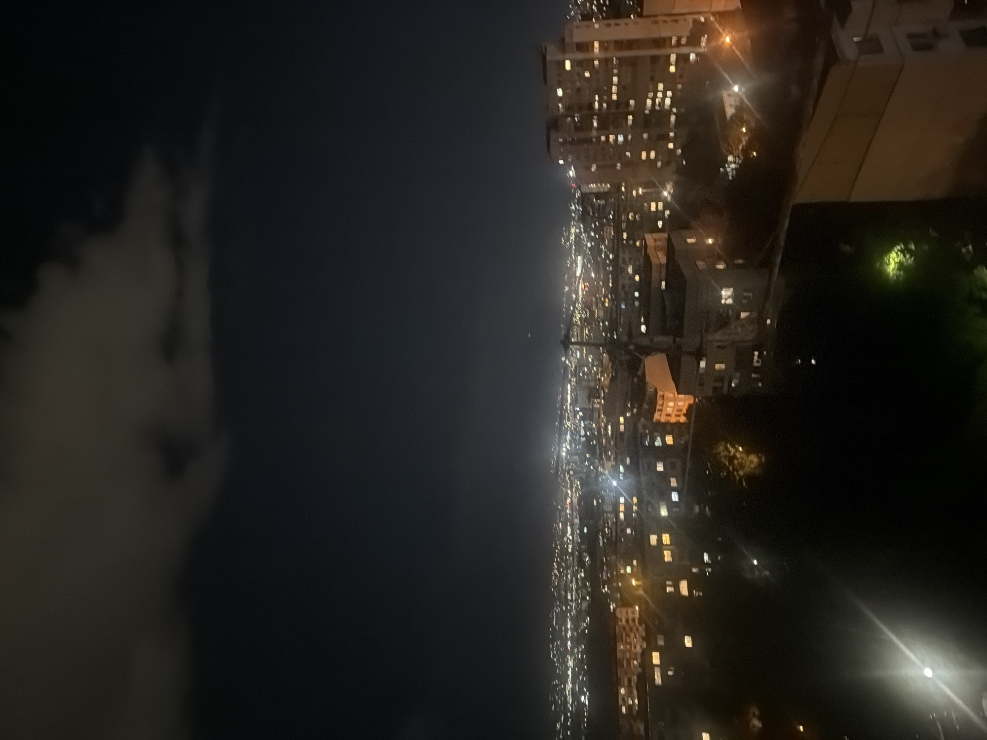 The image presents a panoramic view of a sprawling cityscape at night, captured from an elevated vantage point. The scene is characterized by a dark night sky that occupies the left portion of the frame, with faint, wispy clouds visible against its dark expanse. To the right, a dense urban landscape unfolds, dominated by numerous multi-story buildings, likely apartment blocks or commercial structures. Countless windows in these buildings glow with warm, artificial light, creating a sparkling pattern across the city. The lights extend into the distant horizon, indicating a vast metropolitan area. In the immediate foreground on the right, darker, unlit sections of buildings or landscape, possibly including some trees or green areas, are discernible, along with a bright light source in the lower right corner, possibly a streetlamp or a strong interior light. The time of day is clearly night, with no natural light sources other than ambient light from the distant city. The weather appears calm with some cloud cover. There are no visible people or discernible text within the image. The location context indicates this is a view of Sofia, Bulgaria.