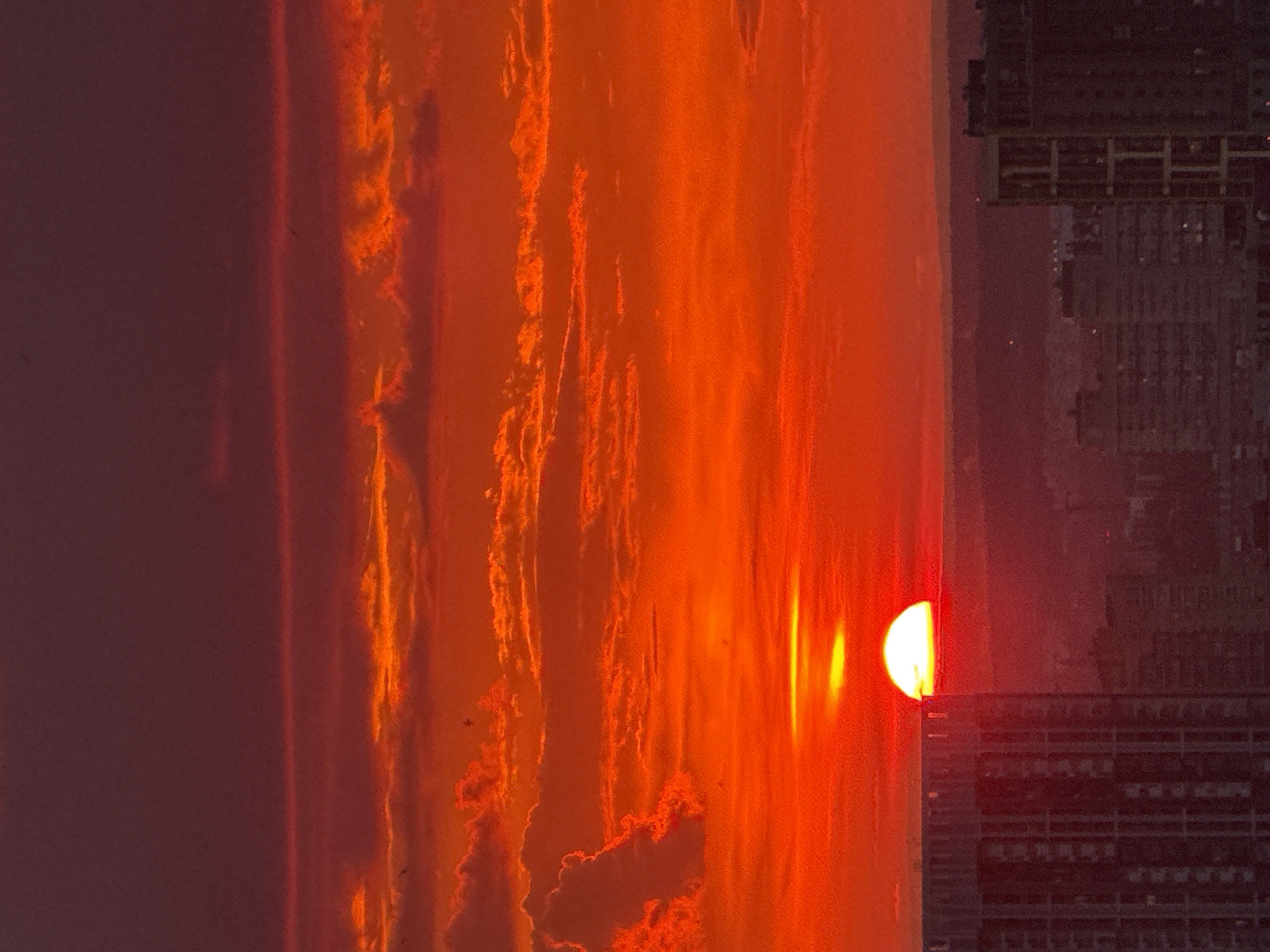 The image captures a vibrant sunset over the city of Savenki, Russia. The sky is ablaze with intense shades of orange and red, with scattered clouds illuminated by the setting sun. The sun itself, a brilliant orb, is partially obscured by the horizon, casting a warm glow. In the foreground, the silhouettes of modern buildings rise against the dramatic sky, indicating an urban setting. The overall scene conveys the dramatic beauty of a sunset in a city environment.