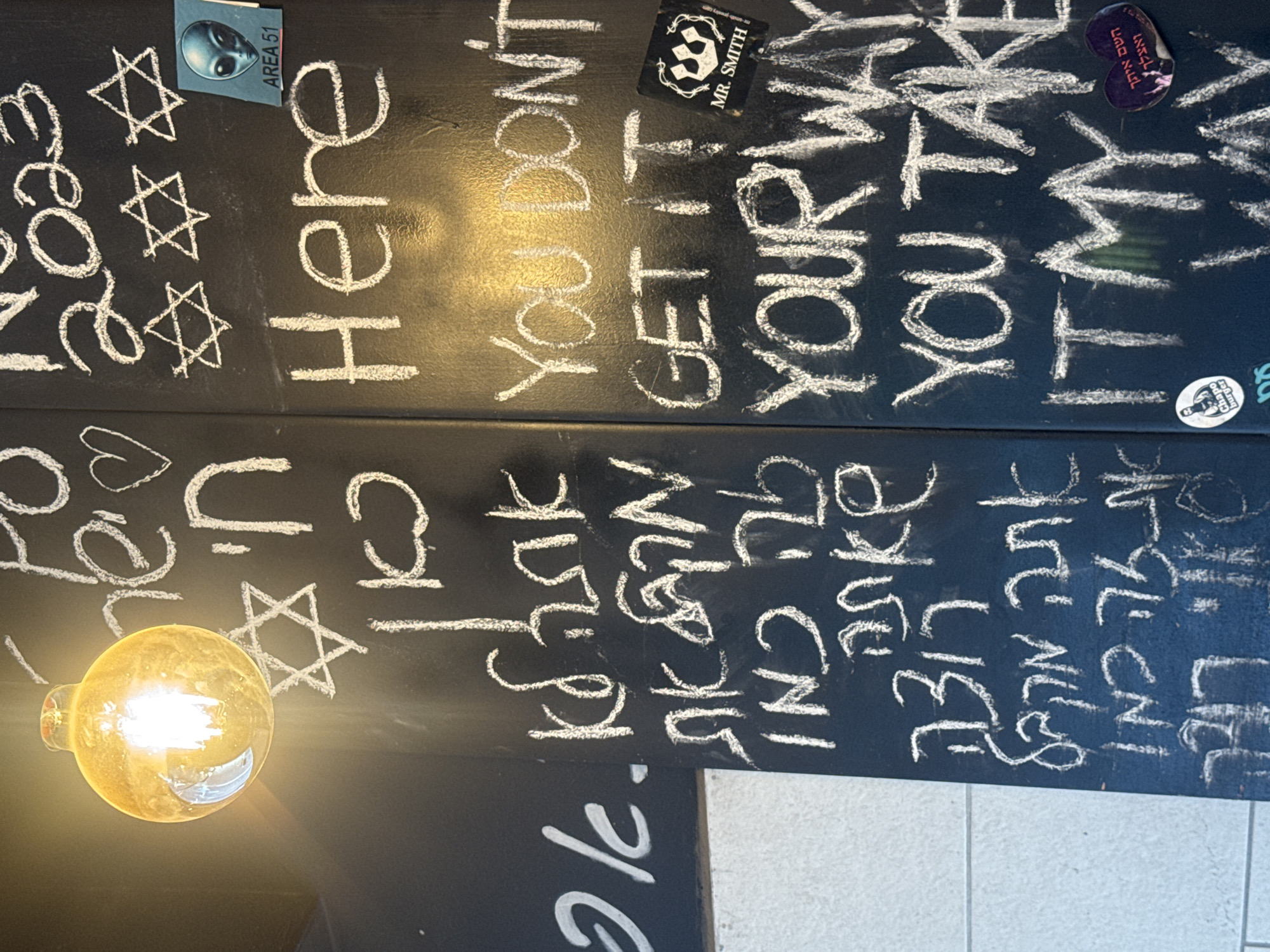 The image shows a blackboard in Ramat Gan, Israel, covered in white chalk writing and stickers. The writing includes English phrases like "YOU DON'T GET IT," "YOUR WAY," "YOU TAKE," and "IT'S MY" interspersed with Hebrew phrases and Jewish symbols (Stars of David). There are also stickers with logos and symbols. A lit lightbulb is positioned on the lower left side of the blackboard.
