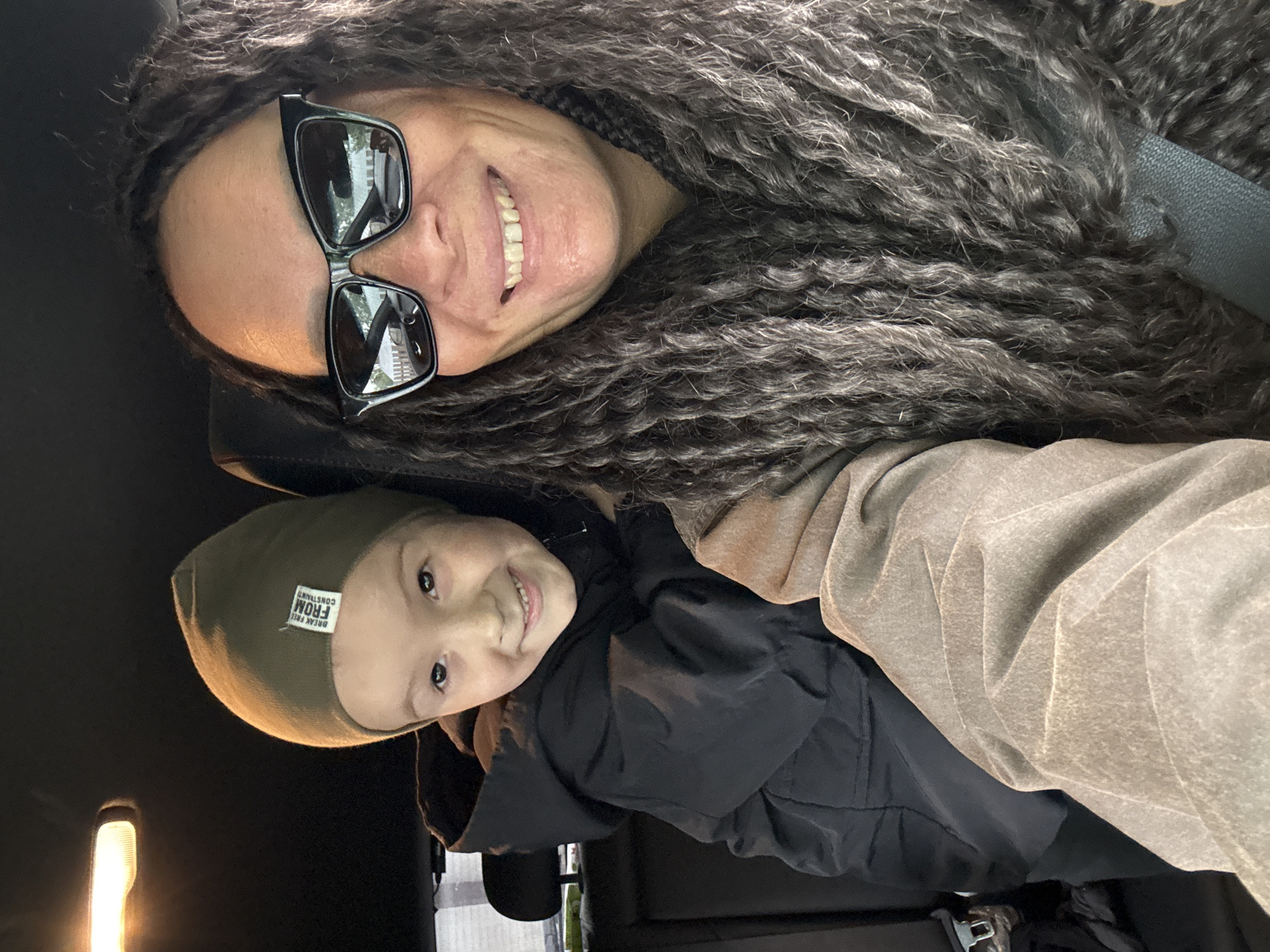 A selfie-style photograph captures an adult woman and a young child smiling inside a vehicle in Chaikovsky, Russia. The image is oriented sideways, with the woman positioned above the child.
The woman, on the left of the image (top in the original orientation), has long, very dark, textured hair, possibly dreadlocks or thick braids, which appear to have grey highlights. She wears black-framed sunglasses that reflect an outdoor scene with trees and buildings. She is smiling broadly, revealing her teeth, and a dark seatbelt crosses her chest.
Below the woman, a young child is also smiling and looking towards the camera, showing teeth. The child wears a dark green or olive-colored beanie hat, on which a white rectangular label with black text is visible, reading "BREAK FREE FROM CONSTRIANT" (the last word appears to be a misspelling of "CONSTRAINT"). The child is also wearing a dark, possibly black, jacket. Next to the child, a light brown or tan fabric is visible, possibly part of their clothing or the car's interior.
The background is mostly dark, suggesting the car's interior. A warm, glowing light source, possibly an interior car light or a reflection, is visible in the bottom left corner. Both subjects appear happy and content, engaged in a casual moment, likely while traveling or stopped.