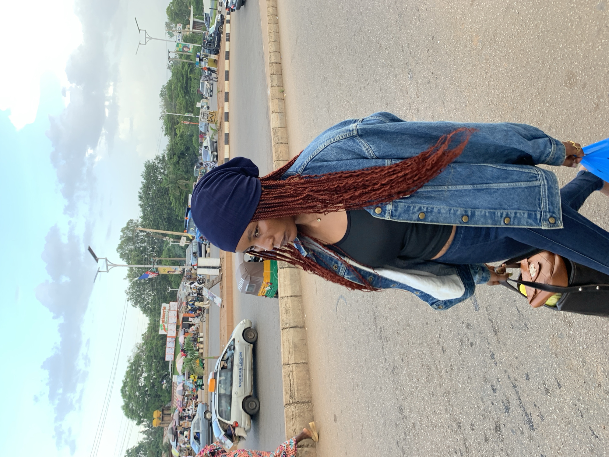 A woman with braided hair and a blue headscarf walks across a street in Jos, Nigeria, carrying a suitcase and a blue plastic bag. She wears a denim jacket over a black tank top and jeans. In the background, a busy street scene unfolds with various vehicles, market stalls with colorful umbrellas, and people milling about. The sky is partly cloudy.