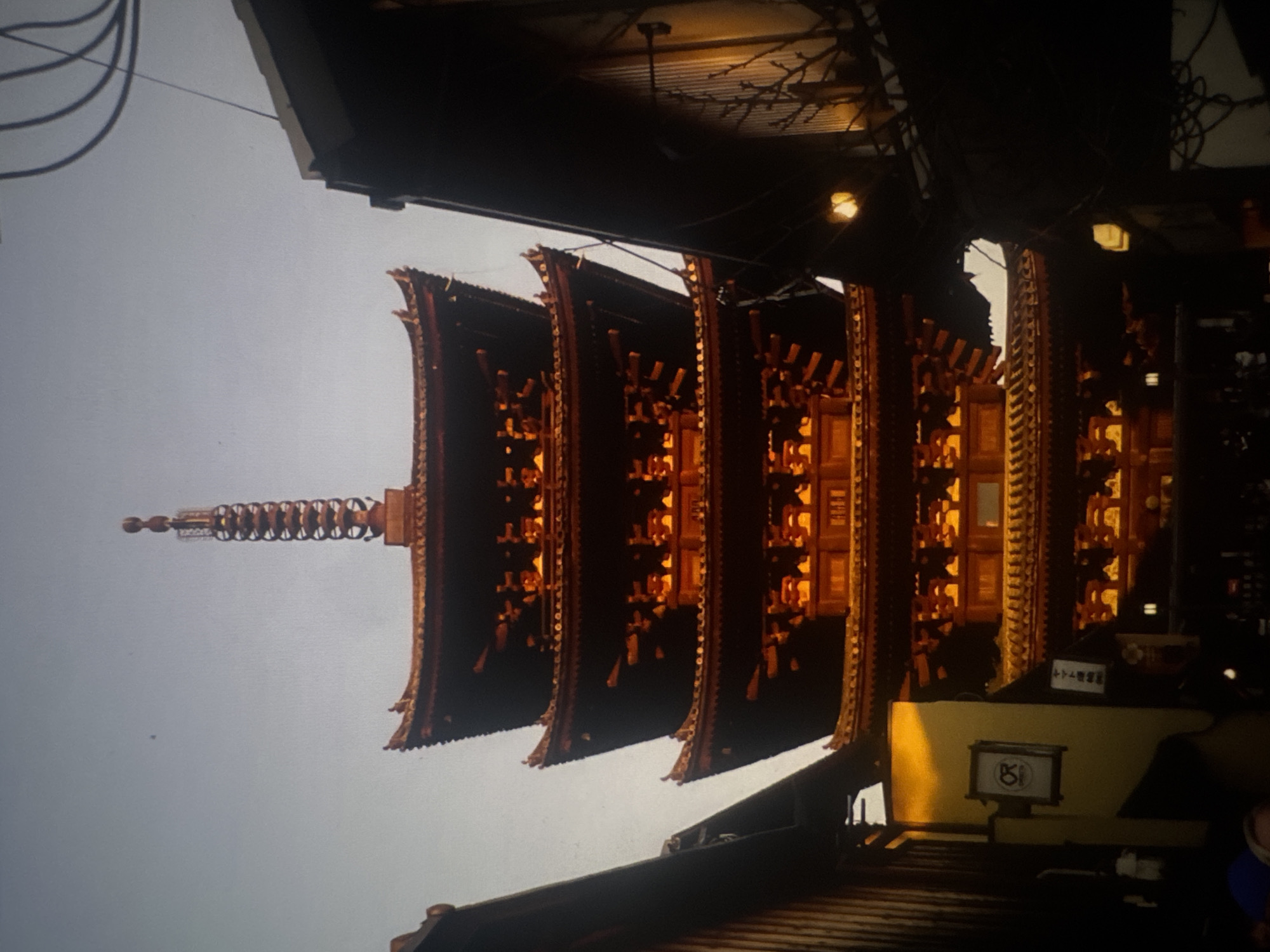 The image shows a multi-story pagoda-style building, likely a temple or shrine, with a distinct tiered roof structure. The building is illuminated from within, casting a warm orange glow through the openings in its facades. The pagoda has a dark, reddish-brown exterior with golden accents on the eaves. Atop the structure is a tall, intricate spire. The building is framed by dark, silhouetted buildings on either side, one of which appears to be a traditional Japanese wooden structure with visible vertical planks and a sloped roof. The sky above is a pale, hazy blue, suggesting either early morning, late evening, or an overcast day. There are some power lines visible in the upper right portion of the image. In the lower left, a portion of a yellow wall is visible with a framed sign containing a logo. The image captures an atmospheric and architectural scene.