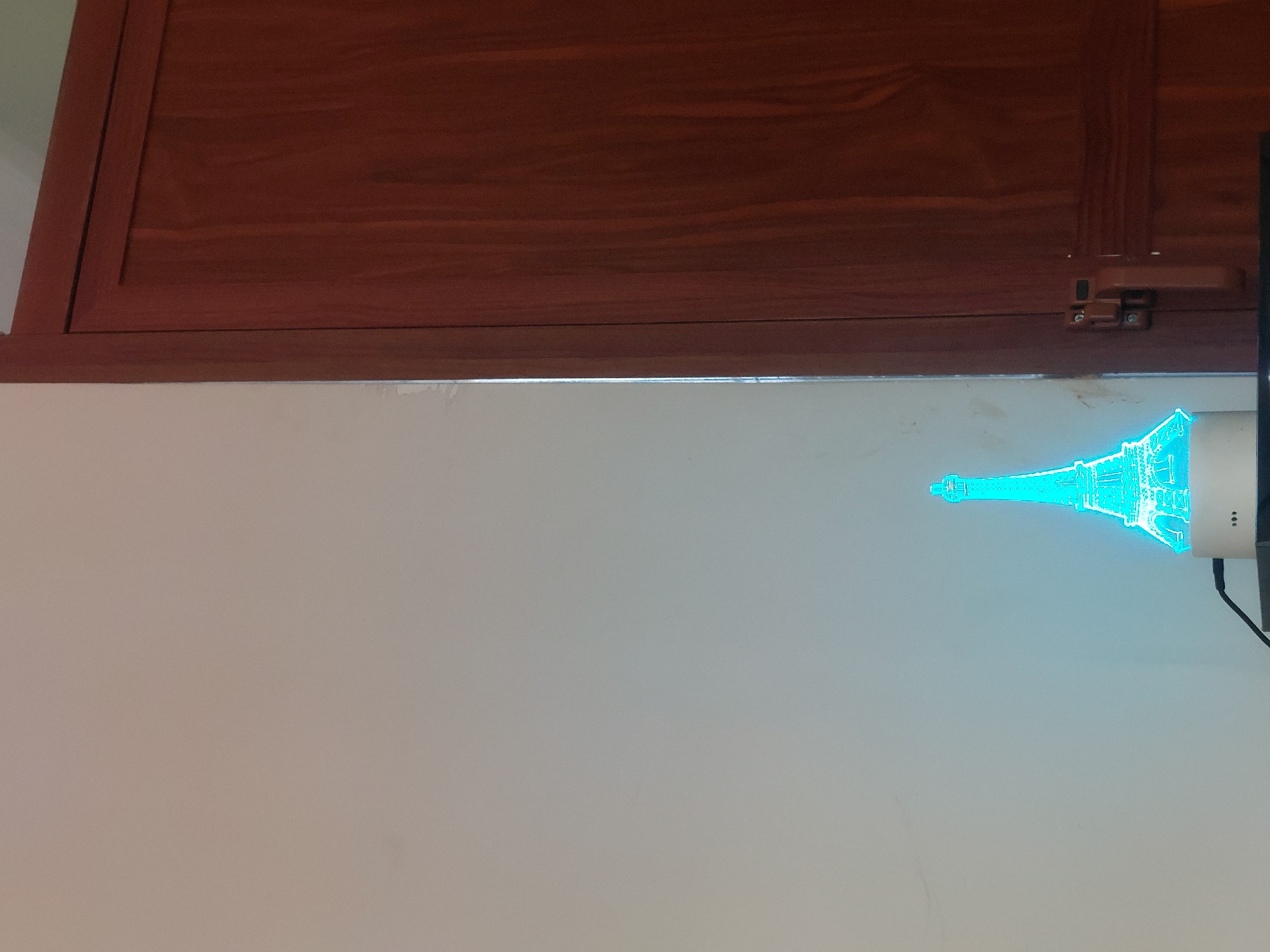 The image shows a wooden cabinet above a light grey wall. On the right side of the wall is a glowing blue Eiffel Tower night light. The scene is indoors. The location of this image is unknown, and there is no indication that it is in Dar es-Salaam, Tanzania.