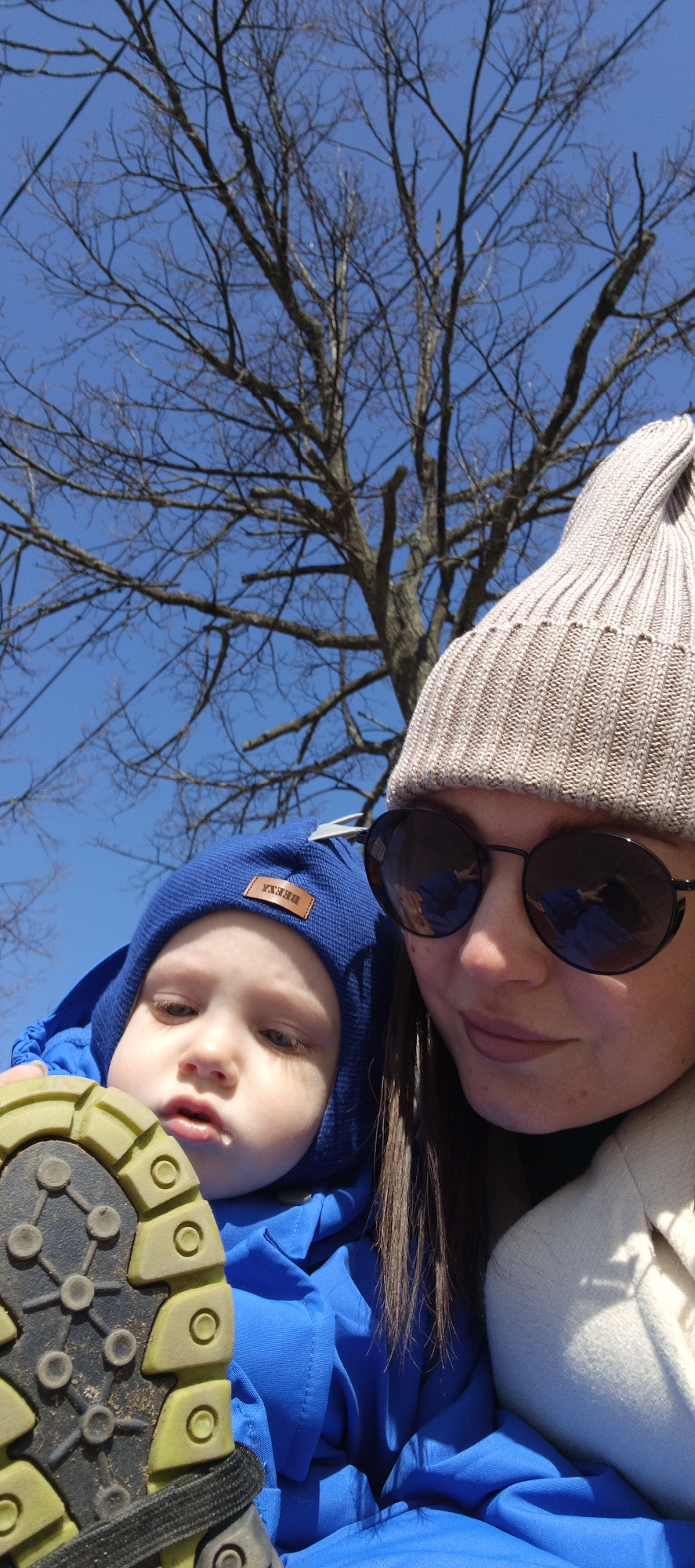 A woman and a toddler are in an outdoor setting, possibly Chaikovsky, Russia. The woman is wearing a tan knit hat and round sunglasses, with dark hair visible beneath. The toddler wears a blue hat and blue jacket. Part of the sole of a child's shoe is visible in the foreground. A bare tree with many branches stands behind them, set against a clear blue sky.