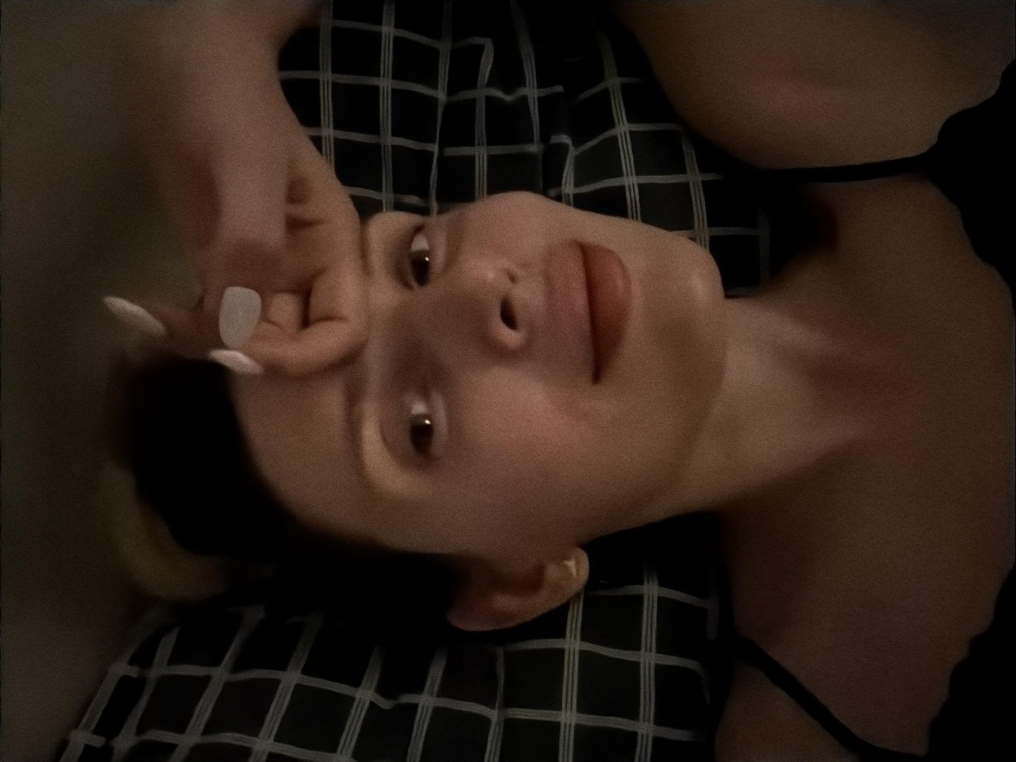 A woman is lying on a black and white checkered surface. She is facing the camera with her right hand touching her forehead. The woman has dark hair, fair skin, and is wearing a black top. The background is out of focus and dark. The location is not discernible from the image.