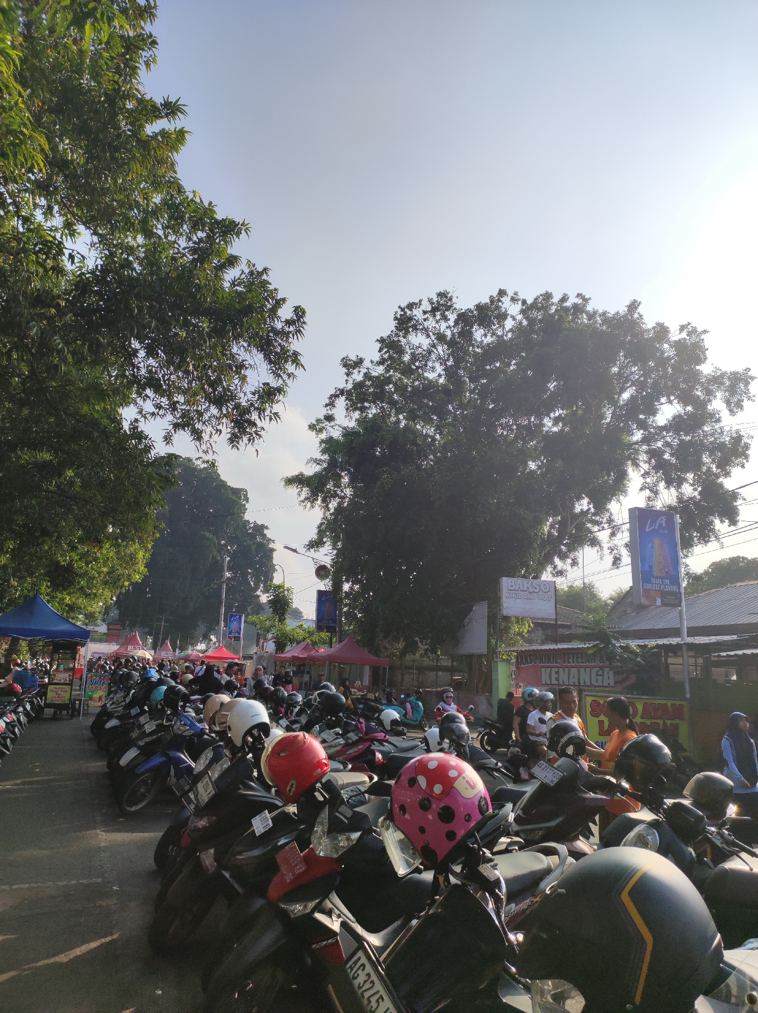 The image depicts a street scene in Blitar, Indonesia, with a large number of parked motorcycles filling the foreground and midground. The motorcycles are neatly arranged in rows, suggesting a designated parking area. Several individuals are visible in the scene, some standing near the motorcycles, while others are walking or conversing. They are dressed in casual attire.
In the background, there are various stalls or small shops set up, indicated by colorful tents and signage. The signage includes text in Indonesian, such as "BARSO KIKIL" and "SO AYAM LAM..." which likely refer to food vendors. A large tree dominates the upper left portion of the image, with its branches extending across the sky. Another large tree is visible in the mid-right background.
The sky is bright and clear, with a hint of haze or lens flare from the sun, which is positioned high and to the right. The overall impression is of a bustling outdoor market or event, possibly on a sunny day, with a significant number of people and their vehicles present. The presence of multiple tents and the variety of food-related signage strongly suggest a local market or street food festival.