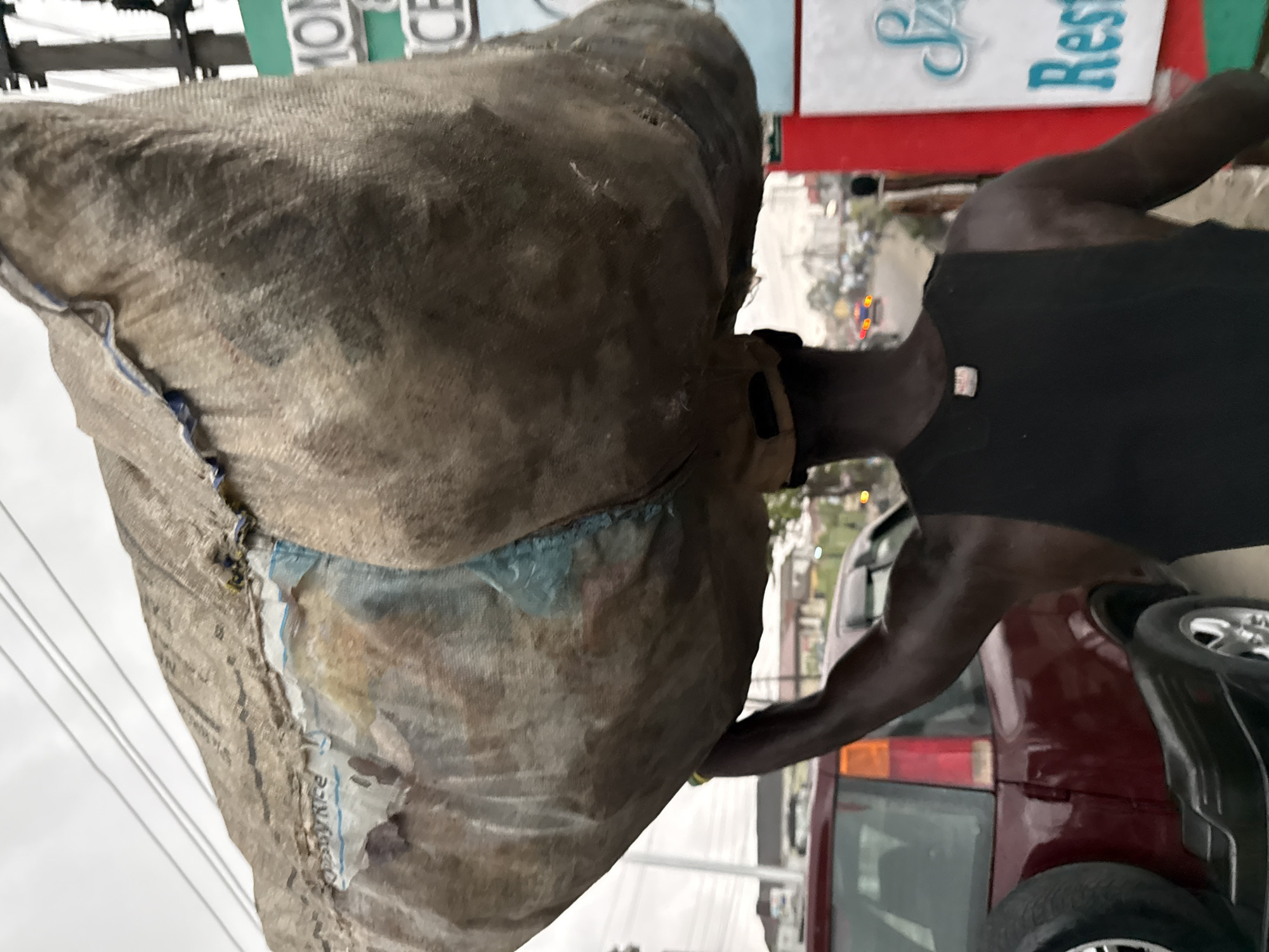 This portrait-oriented image depicts a person carrying a very large, bulky sack on their head and left shoulder, likely in a street environment in Ado, Nigeria. The individual appears to be a dark-skinned male, wearing a black sleeveless shirt and a cap, with visible muscular arms.
The sack, made of a light brown, coarse, woven material, is immense and appears heavily loaded, almost engulfing the person's upper body. Through a torn section of the sack, a blue plastic inner bag or lining is visible, bearing the partially legible text "OMMY RICE", indicating the contents are likely bags of rice.
In the background, a maroon or dark red car is partially visible on the right, suggesting a street setting with traffic or parked vehicles. Above, fragments of street signs can be seen: a green sign with white text, partially reading "MON", and a white sign with blue text that seems to say "Sjz Rest", possibly part of a restaurant sign. The sky is overcast, indicating a cloudy day. The scene captures an individual engaged in manual labor, transporting goods.