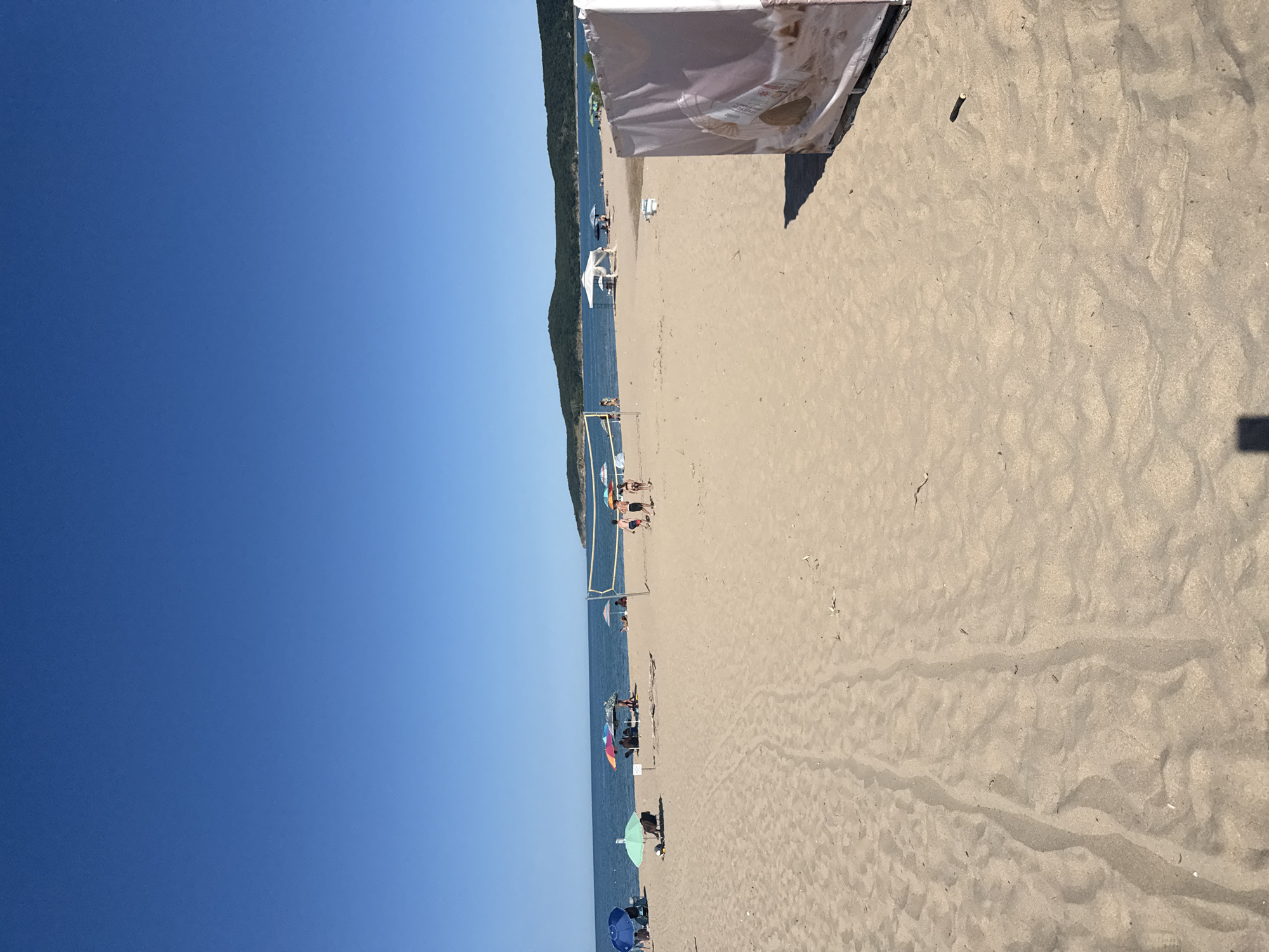 The image depicts a sandy beach scene in Dyuni, Bulgaria, under a clear blue sky. In the foreground, tire tracks are visible in the sand. Further back, several beach umbrellas are set up, with a few people seated or standing beneath them. A volleyball net spans across the sand, and a group of people are playing volleyball. The sea is visible in the background, with a gently sloping, tree-covered hill rising from the coastline. A white canvas structure is positioned in the right foreground of the image.