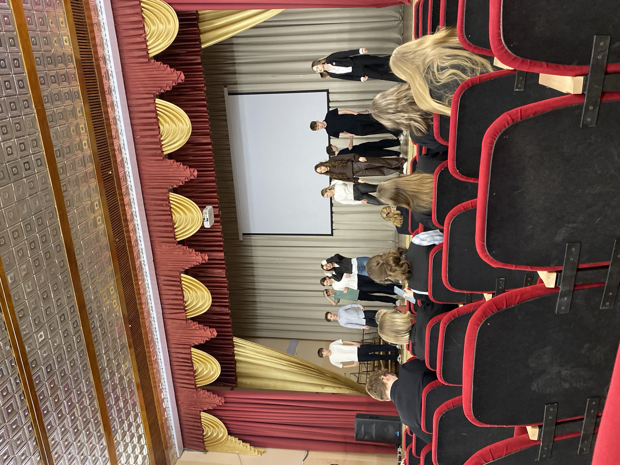 The image depicts an indoor scene within what appears to be an auditorium or a school hall, viewed from the audience seating area looking towards a stage.
On the stage, a group of approximately ten to twelve young individuals, likely teenagers or young adults, are standing in two rows. They are dressed in a mix of casual and semi-formal clothing, including dark suits, white shirts, and various colored tops and trousers. Their posture suggests they are either performing, presenting, or rehearsing. A large white projection screen is prominently positioned on the left side of the stage, with a projector visible near its base. The stage is adorned with elaborate red and gold/cream curtains. A black speaker is situated on the floor at the front left of the stage.
In the foreground and extending into the midground, several rows of red and black audience seats are visible. These seats feature black upholstery with red piping and wooden armrests. The backs of several audience members' heads and shoulders are discernible, facing the stage. Many appear to have light-colored hair. The ceiling above the audience features ornate, patterned panels. The lighting is consistent with an indoor setting, providing clear visibility throughout the space. There is no visible text in the image. The scene captures a performance or presentation in progress.