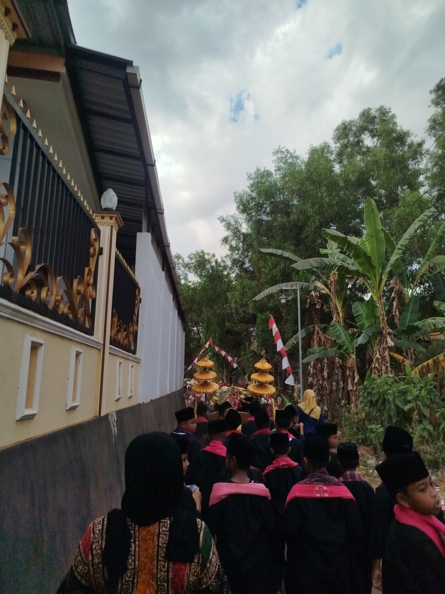In Parseh, Indonesia, a procession of young people, most wearing black skullcaps and robes with red trim, walks along a paved pathway. A woman in a yellow hijab is visible further back. The group is moving past a building with a light yellow facade and a dark metal roof supported by exposed beams, bordered by a black and gold ornate fence. To the right of the building and pathway, lush green trees, including banana plants, are abundant. Two red and white striped flags are planted near decorative, multi-tiered golden structures, suggesting a cultural or religious event. The sky is overcast with scattered clouds.