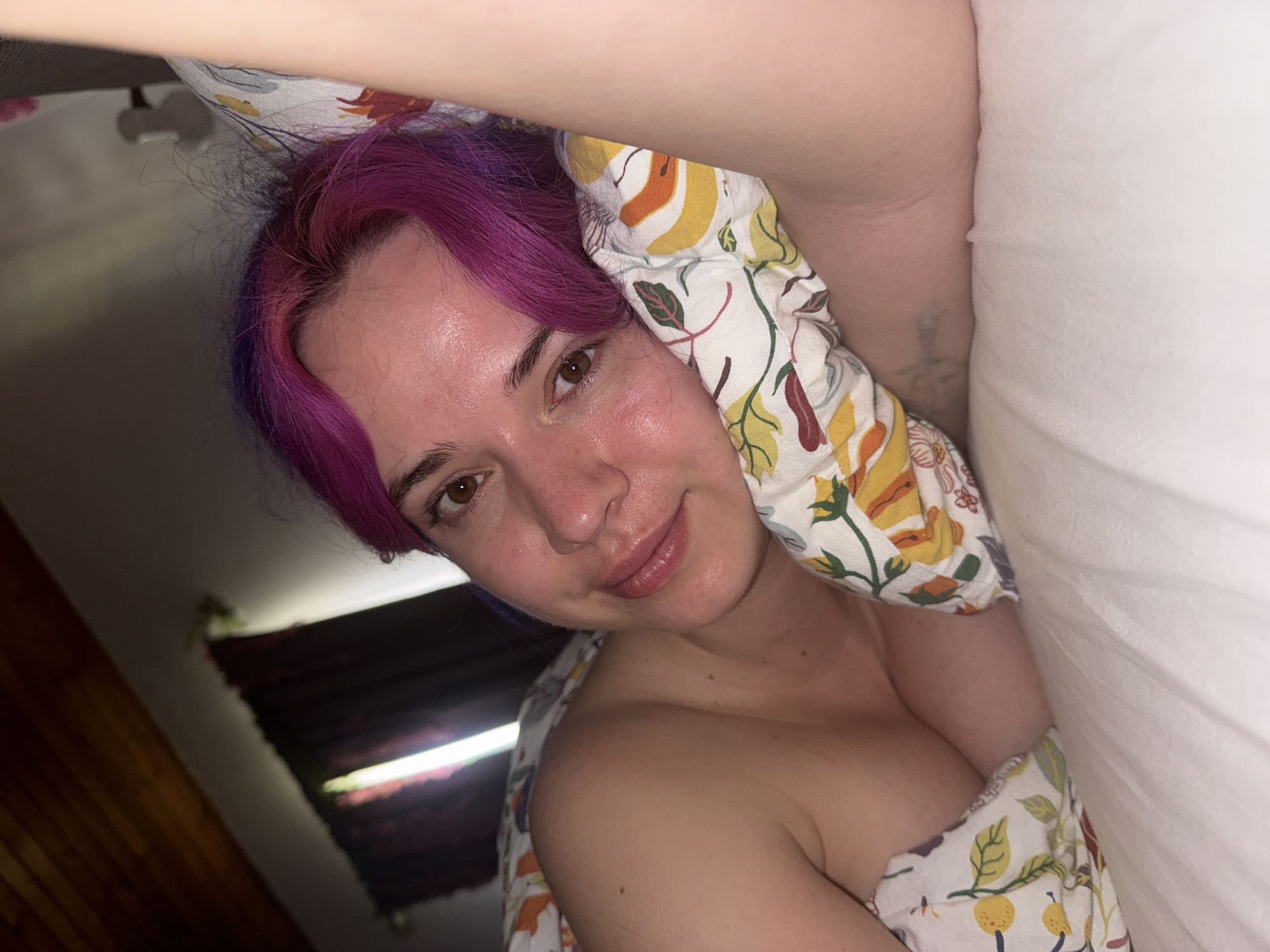 A woman with purple hair is lying in a bed with white sheets and a floral-patterned pillow and blanket. The photo appears to have been taken indoors. There is a window with closed blinds visible in the background. The location in the city of City of Zagreb, Croatia, cannot be determined from this image.