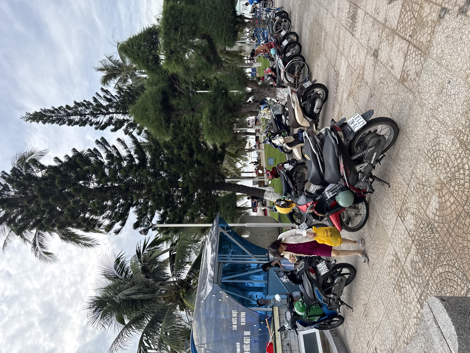 The image captures a street scene in Nha Trang, Vietnam, likely near the beach, given the presence of palm trees and the general tropical atmosphere. In the foreground, a row of parked motorcycles occupies a tiled pedestrian area. A woman in a dark pink jumpsuit and a white cardigan walks towards the right, carrying a bright yellow shopping bag. Her sandals are visible as she walks on the patterned tiles. Behind her, several other motorcycles are parked, some with helmets on them, including a blue Grab-branded delivery motorcycle. To the left, a modern, transparent blue structure with a curved roof stands, possibly a public restroom or information kiosk, with text visible on its side in Vietnamese. Two young women are standing near the entrance of this structure. Further into the background, tall pine trees and more palm trees line the area, with a glimpse of a beach and the ocean in the far distance. Several people are casually strolling on the beach and near the water. The sky is overcast with scattered clouds. The scene appears to be a public space designed for pedestrians and parking, with amenities and natural elements creating a coastal city ambiance.