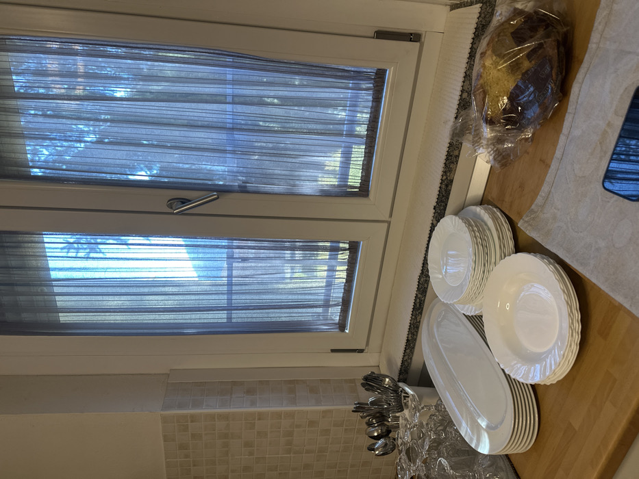 The image captures an indoor kitchen scene, focusing on a window and a countertop. A white-framed window with two panes dominates the left side of the frame. Each pane is covered by a sheer, dark grey blind or curtain with horizontal stripes, allowing diffuse light and glimpses of green foliage outside, suggesting a bright, sunny daytime. A silver-colored window handle is visible on the upper pane.
On the right side of the image, a light wooden countertop holds various items. Multiple stacks of white dinner plates, white bowls, and a large oval white serving platter are neatly arranged. These dishes feature a subtle textured or fluted rim. Adjacent to the plates, a clear, possibly crystal, container holds an assortment of silver-colored cutlery, including spoons and forks. A loaf of dark bread, encased in clear plastic packaging, rests on a light-colored, patterned fabric cloth or towel. A dark-colored smartphone, reflecting light, is partially visible near the bread. Below the window, a light-colored tiled backsplash adds texture to the wall. No people are visible, and no specific activity is actively occurring, though the setup of dishes and bread implies preparation for a meal or gathering. There is no visible text.