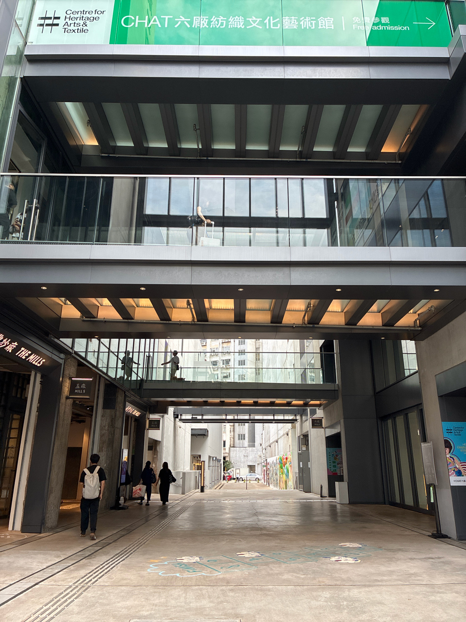 This is a street-level, eye-level, full shot of a modern urban environment in Hong Kong, China. The scene depicts an open-air walkway between two multi-story buildings. The architecture features extensive use of glass, steel, and concrete. Multiple elevated walkways or bridges connect the buildings overhead, with people visible on them. The walkway on the ground level has distinct parallel lines embedded in the pavement, possibly indicating former tram tracks or a design element. There are also colorful chalk drawings on the ground, resembling hopscotch patterns.
In the foreground, on the left side of the walkway, a young man wearing a white backpack and dark clothing is walking away from the camera. Further down the walkway, two women, also walking away from the camera, are visible. One is carrying a shopping bag.
The buildings themselves display signage, including English and Chinese characters. One prominent sign at the top reads "Centre for Heritage Arts & Textile" and "CHAT 六廠紡織文化藝術館 | 免費參觀 Free admission" in green and white. Other signs indicate "THE MILLS".
The overall impression is one of a contemporary, lively urban space, likely a commercial or cultural district, designed with a blend of modern aesthetics and hints of historical context, suggested by the "heritage" and "textile" elements in the signage. The lighting suggests it is daytime, with natural light filtering through the open spaces and glass structures.