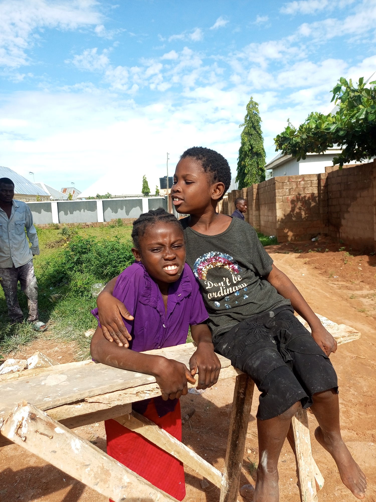 The image depicts two young children, a boy and a girl, sitting on a makeshift wooden bench outdoors. The boy, wearing a dark t-shirt with a colorful sequined design and black shorts, is positioned on the right, his arm around the girl. The girl, wearing a purple shirt and a red garment beneath, is seated on the left, her face turned slightly towards the viewer with a strained expression, baring her teeth. They are in an outdoor setting with a dirt path in the foreground and a grassy area with some vegetation behind them. In the background, a man in a light blue shirt and gray pants is visible walking away from the camera. A brick wall and some buildings are also present in the background, suggesting a semi-urban or rural environment. The sky is blue with scattered white clouds. The scene appears to be set in Tempure, Nigeria.