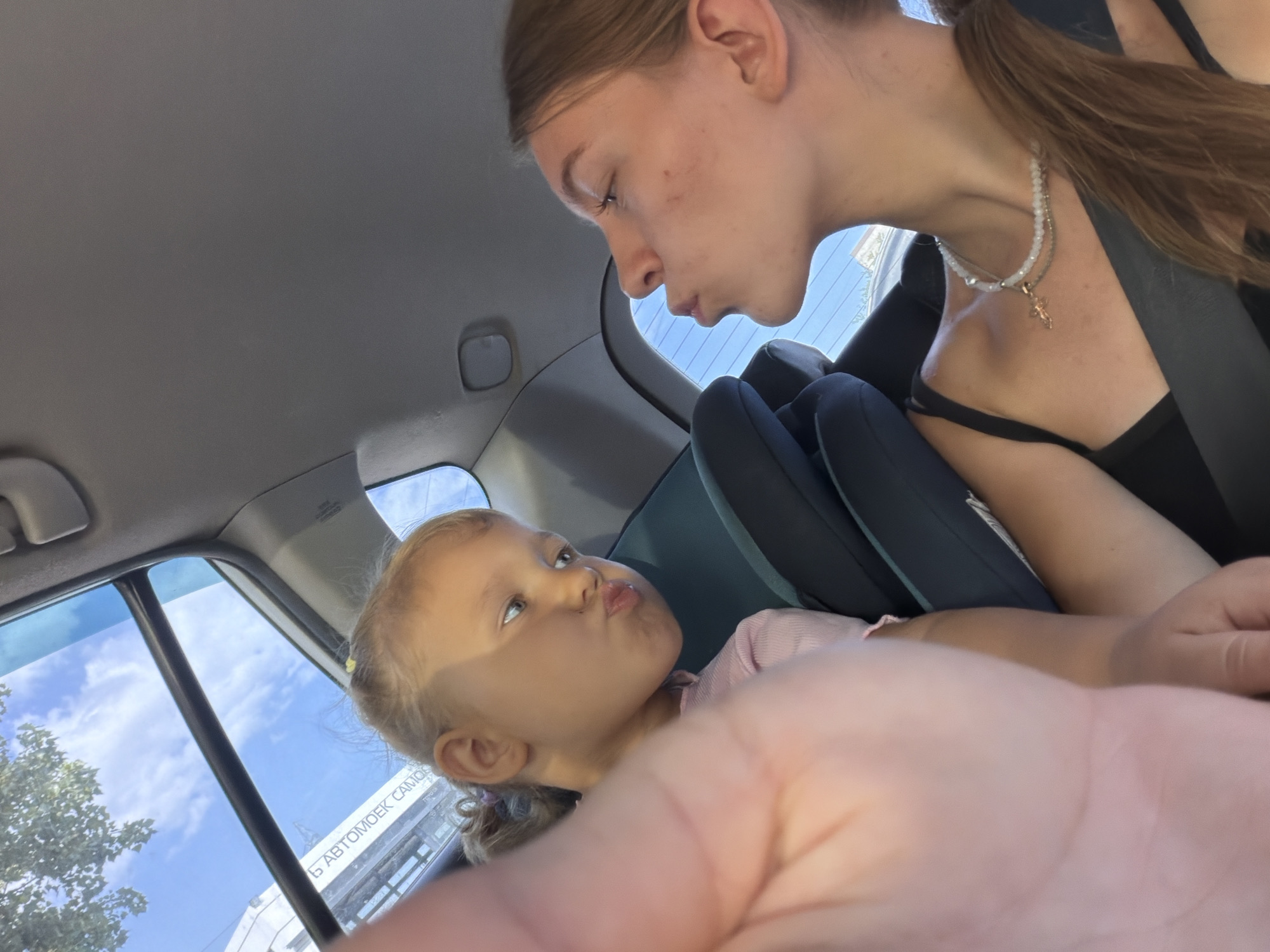 The image shows two females in the interior of a car, likely a passenger vehicle. A young child, appearing to be a toddler, is seated in a car seat and looking upwards with pursed lips. The child has blonde hair tied in pigtails. An older female, with blonde hair pulled back in a ponytail, is leaning in towards the child, appearing to be either speaking to or playing with the child. She is wearing a black top and a necklace. The car's interior is visible, including the headliner, car seat fabric, and window. Through the window, a blue sky with clouds and some trees are visible, along with a sign with Cyrillic text that appears to read "Автомобил самообслуживания" (Car self-service). This sign, combined with the style of the car and the overall environment, suggests the location is Rostov-on-Don, Russia. The perspective of the photo, taken from a low angle and partially obscured by a hand in the foreground, suggests it was taken by one of the occupants of the car.