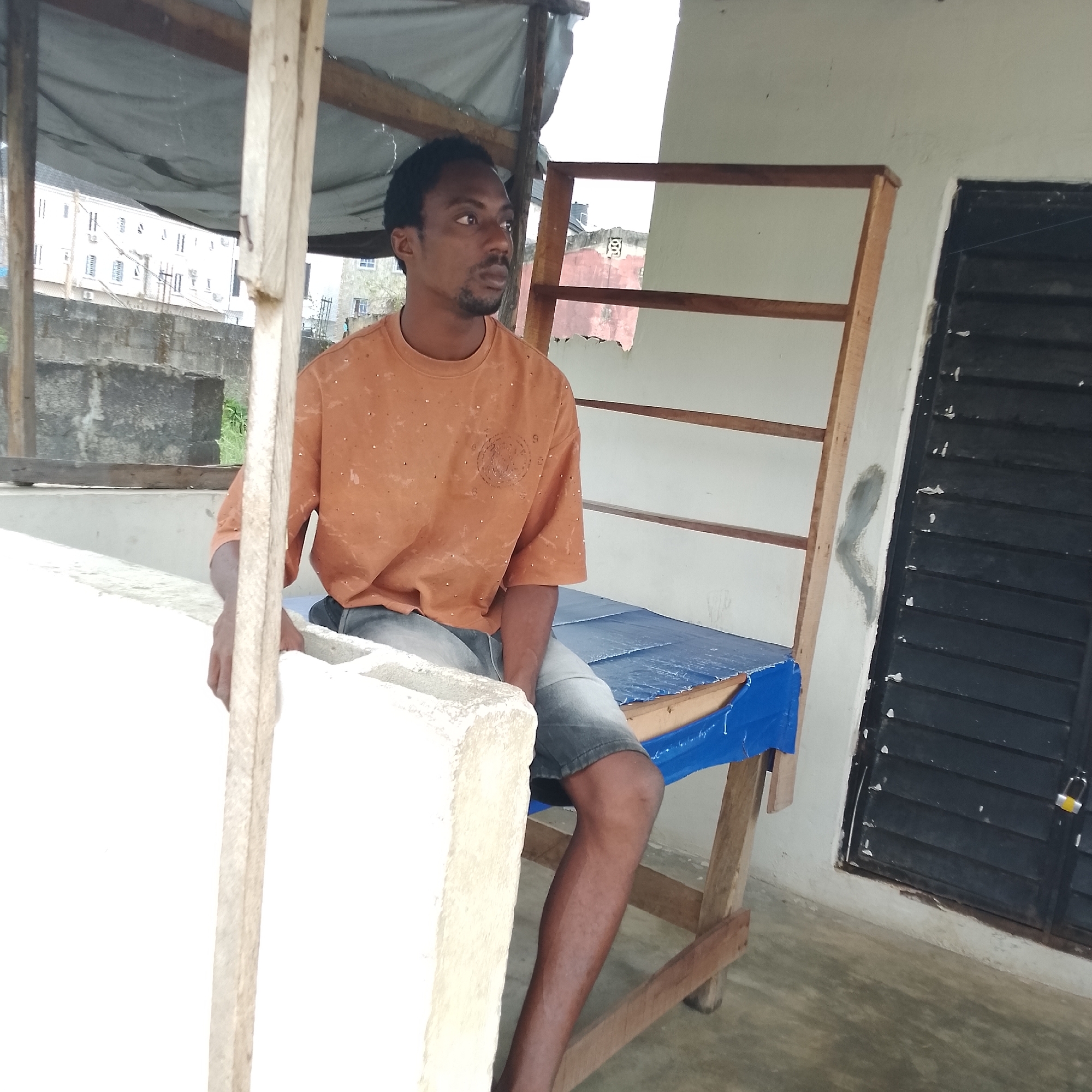 In Ado, Nigeria, a man is seated under an open-air canopy. He is looking to his right and appears to be in his late 20s or early 30s. He is wearing an orange t-shirt with small silver embellishments and denim shorts. The man is seated on a rough wooden structure which has a blue tarp stretched across it, possibly serving as a makeshift bed or bench. The background reveals a textured wall with a barred window and in the distance, buildings and a concrete wall. The lighting suggests it is daytime.