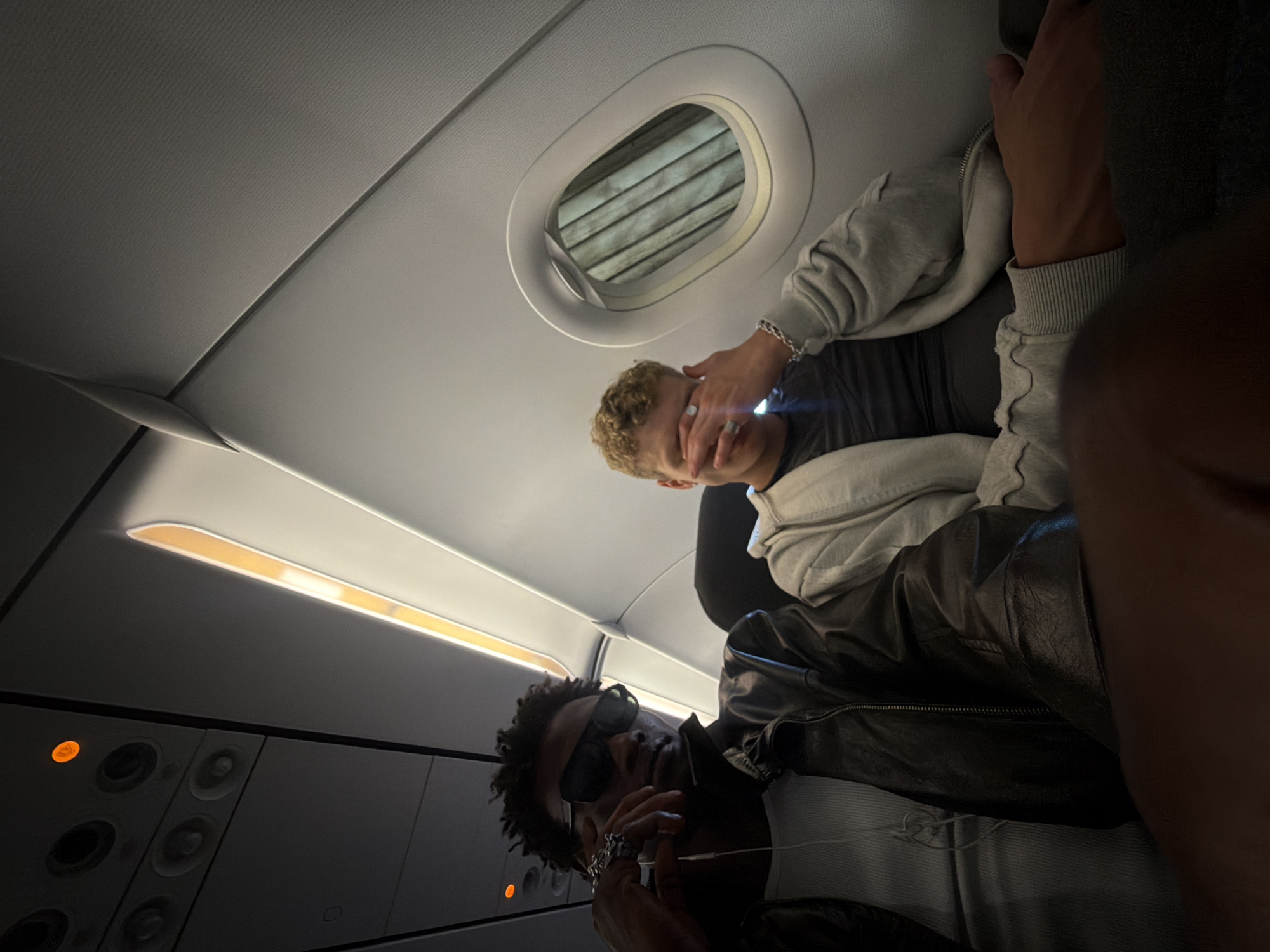 The image shows two young men seated on an airplane, possibly en route to or from Crawley, United Kingdom. The man in the foreground, on the left, wears a black leather jacket over a white tank top and dark sunglasses. He is wearing a large, ornate ring on his right hand. The man to his right, with blond curly hair, wears a grey hooded sweatshirt and has his hand covering his eyes. The interior of the plane is visible, with overhead compartments and an airplane window with blinds. An orange light is illuminated above the men, indicating a possible flight status or warning. The overall lighting suggests a dim cabin environment.