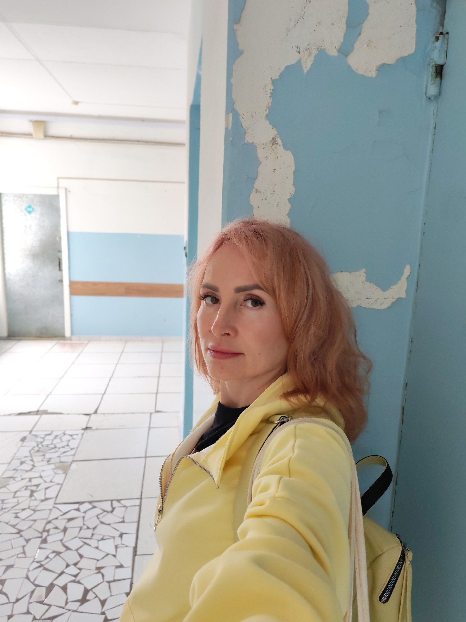 A woman with reddish-blonde hair, wearing a yellow hooded jacket and a backpack, stands in a hallway. The hallway has light blue walls with peeling paint and white trim. The floor is tiled with a mosaic pattern of small white tiles. In the background, a door is visible on the left, and a wooden handrail runs along the wall. The lighting is consistent with indoor lighting.