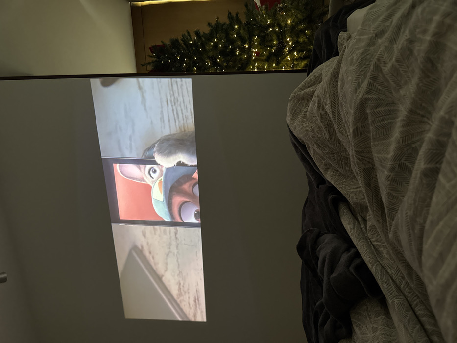The image displays an indoor residential scene, likely a bedroom, captured from a low perspective, seemingly from a bed. A wide-format animated film clip is projected onto a light-colored wall in the upper central area. The projection features characters from Disney's "Zootopia"; specifically, Judy Hopps, identifiable by her police cap, is looking over what appears to be a tablet screen, with her furry paw visible to the right and the eyes of another character (likely Nick Wilde) visible beneath. A blurred, reflective object is also visible on the left side of the projected image.
The foreground is occupied by a bed covered with rumpled bedding. A dark grey fleece blanket lies on the left, while a lighter beige-grey blanket with a subtle patterned texture covers the right and central portions. No individuals are present in the frame.
To the right side of the scene, beyond a distinct vertical wall partition, a decorated Christmas tree is visible. The tree is adorned with numerous warm white string lights and some dark ornaments, set against a muted yellow wall. The overall ambient lighting in the room is dim, primarily emanating from the projected image and the Christmas tree lights.
The specific location within the city of Warsaw, Poland, is not discernible from the visual information provided in the image.