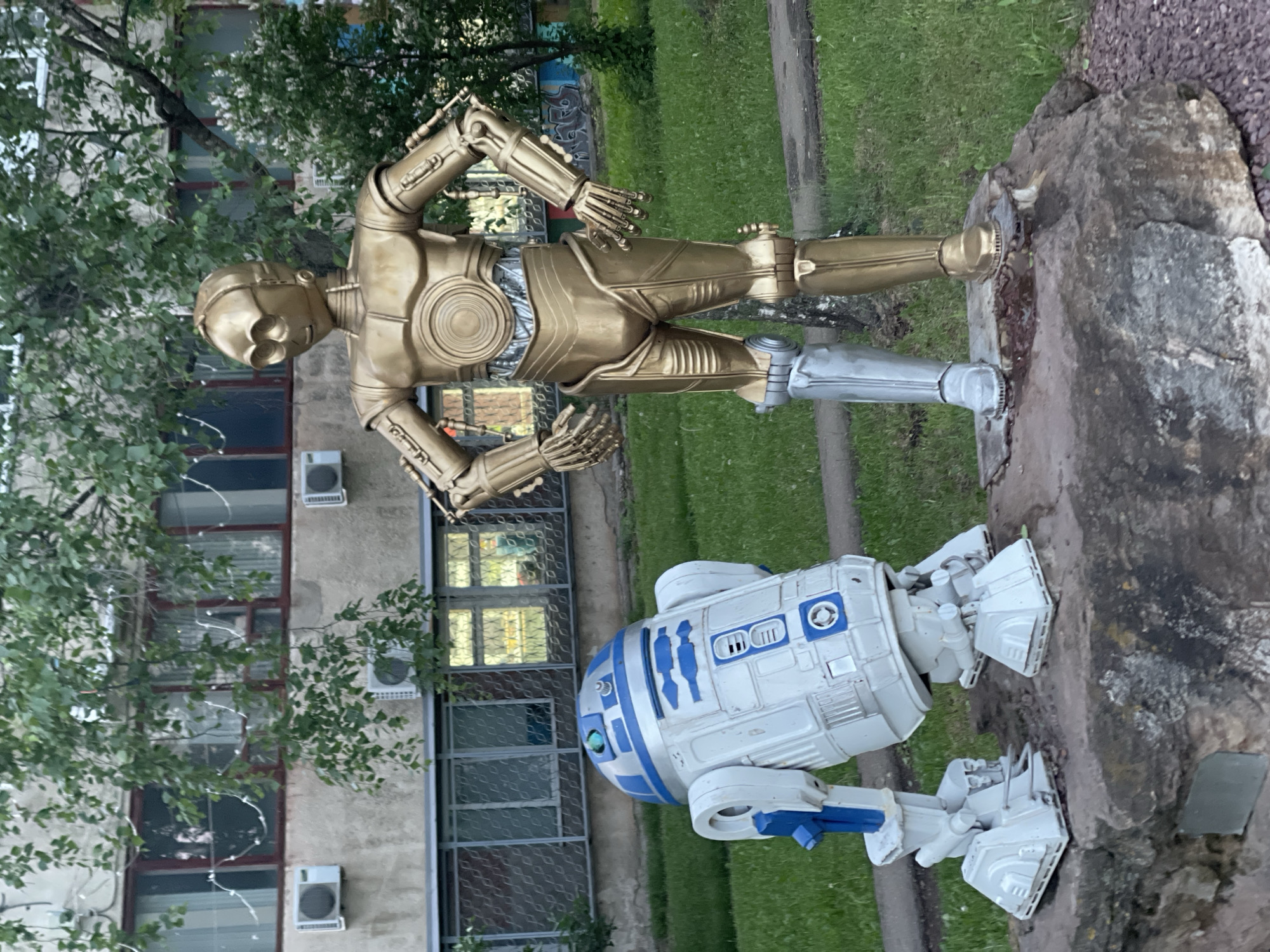 The image shows statues of the Star Wars characters C-3PO and R2-D2 in the city of Izhevsk, Russia. C-3PO is gold colored with a silver lower leg and is positioned lying diagonally on a rock. R2-D2 is white with blue markings and is positioned next to the rock. Behind the statues is a building with many windows and trees. In the background, there is a green grassy area and a sidewalk.