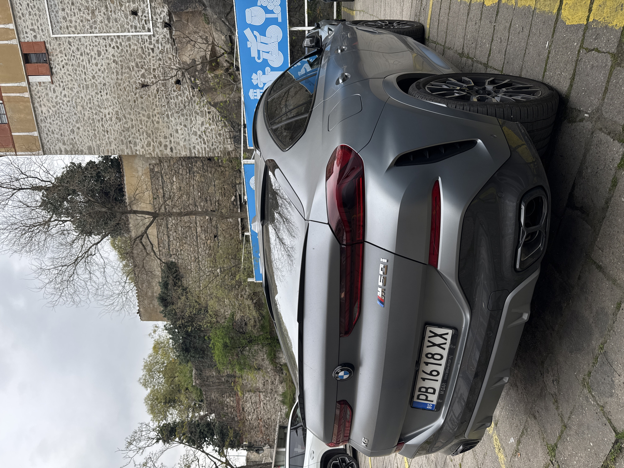 The image shows the rear of a gray BMW X6 M50i parked on a cobblestone or concrete road with yellow painted lines indicating parking spaces. The car has Bulgarian license plates reading "PB 1618 XX." The background features a stone wall and a hill with trees and some buildings, along with a blue sign with white writing and symbols, likely indicating a local attraction or restaurant. Based on the architectural style and general appearance, the location is likely in or near the old town of Plovdiv, Bulgaria. There are no people visible in the image. Another vehicle is partially visible in front of the BMW.