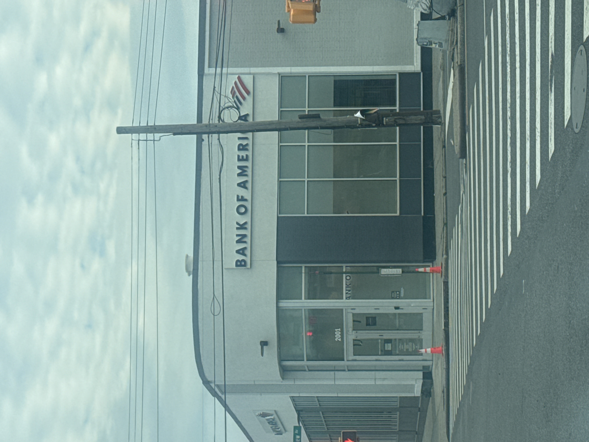 The image shows a Bank of America building with its name visible on the exterior. The bank is located in New York, United States of America. There are several windows on the building, some of which are covered with dark material. A street with a pedestrian crossing is visible in front of the bank, along with two orange traffic cones. A wooden pole with electrical wires runs across the foreground of the image, partially obscuring the building. In the upper part of the building, a yellow traffic signal is visible. The sky is cloudy, suggesting overcast weather. The time of day is unclear. No people are visible in the image.