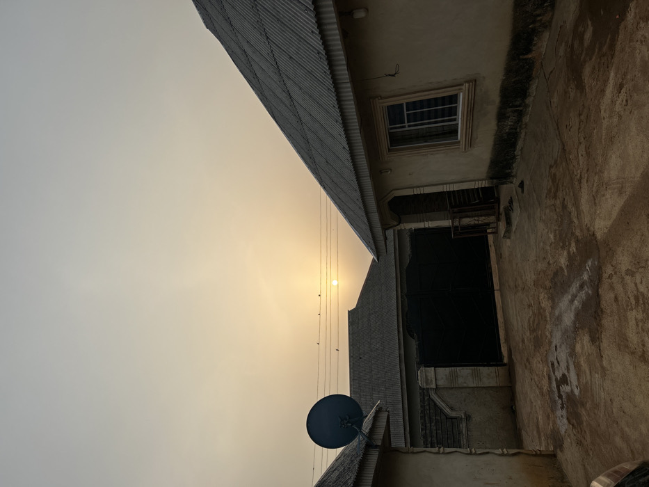A sideways view shows a sunlit sky beside a collection of buildings. Corrugated metal roofs are visible, along with a satellite dish attached to one roof. An adjacent building displays a window with bars and a dark doorway. A concrete wall with dark stains lines the right side of the image. Wires run through the sky, with birds perched on them near the sun.