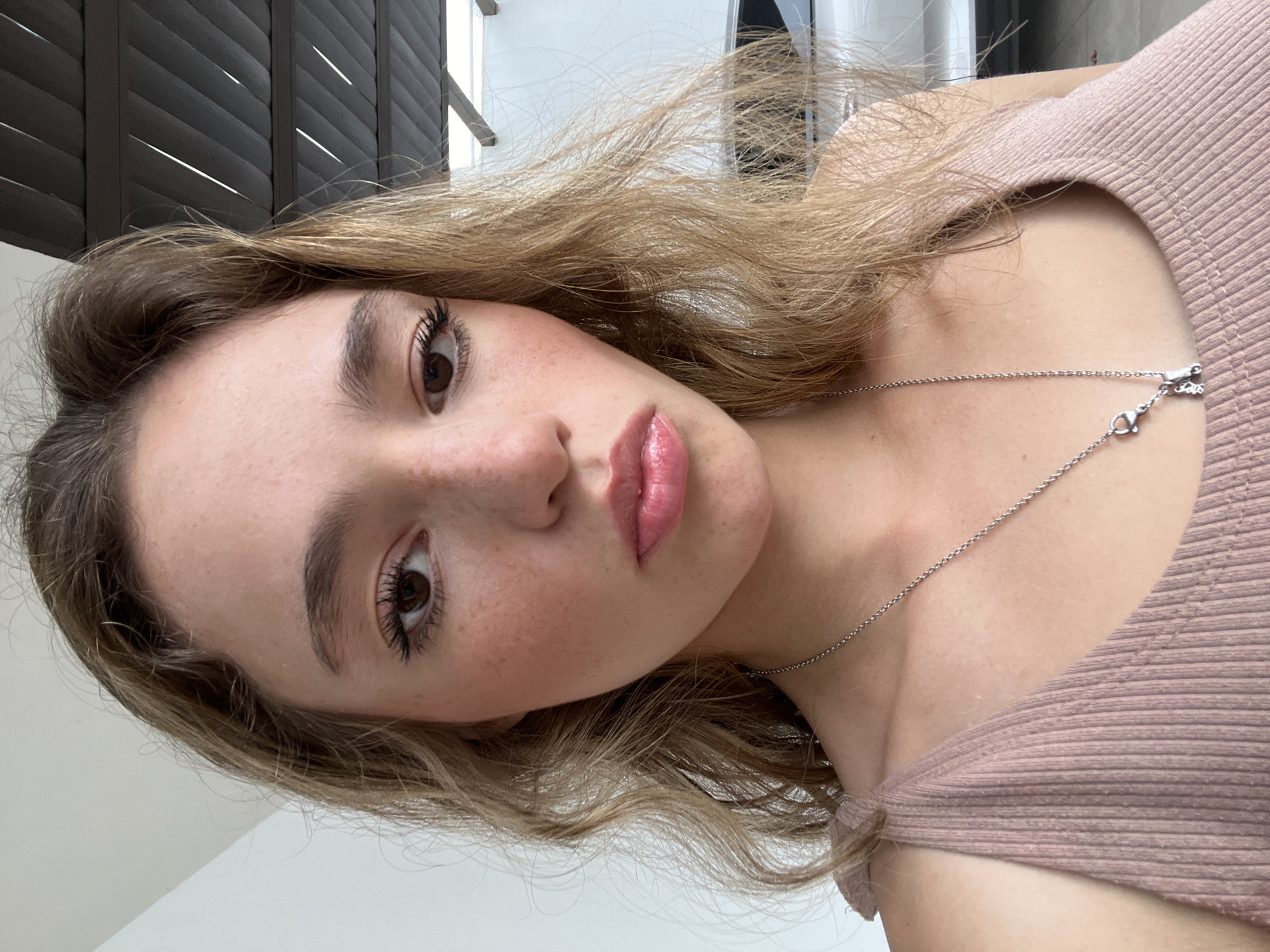 A young woman with light brown hair is centered in the frame, facing slightly towards the camera. She has light skin, brown eyes, full lips with glossy pink lip color, and visible freckles. She is wearing a light pink top and a delicate silver necklace. The background shows a partial view of a dark brown shutter, possibly of a window, and a section of white wall. The image is taken from a slightly elevated angle. The location is indeterminate due to a lack of identifiable landmarks.