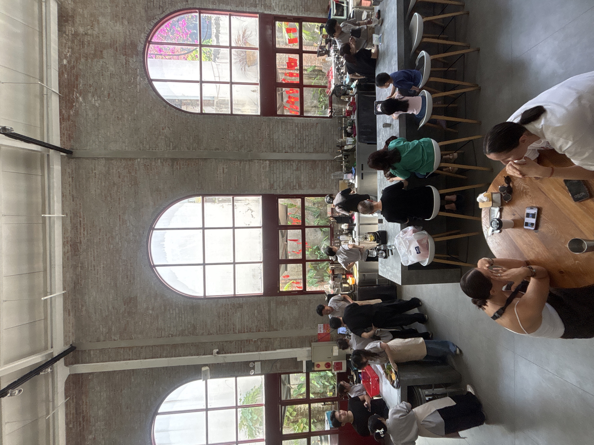 The image depicts the interior of a bright, airy cafe with high ceilings, exposed brick walls, and large arched windows. The cafe appears to be located in Da Nang, Vietnam, given the presence of red lanterns often displayed during Tết celebrations. The space is modern and industrial, with concrete floors and a long counter area where baristas are preparing drinks and food.
Several patrons are seated at the counter, including a family with two young children. Other individuals are standing, some interacting with staff and others looking at their phones. A woman in a white tank top is seated at a round wooden table in the foreground, resting her chin on her hand. A barista in a black uniform is behind the counter, preparing a drink.
The cafe is well-lit, with natural light streaming in through the windows. There are plants visible both inside and outside the cafe, adding a touch of greenery to the space. The overall atmosphere is casual and relaxed, with people enjoying their time at the cafe.