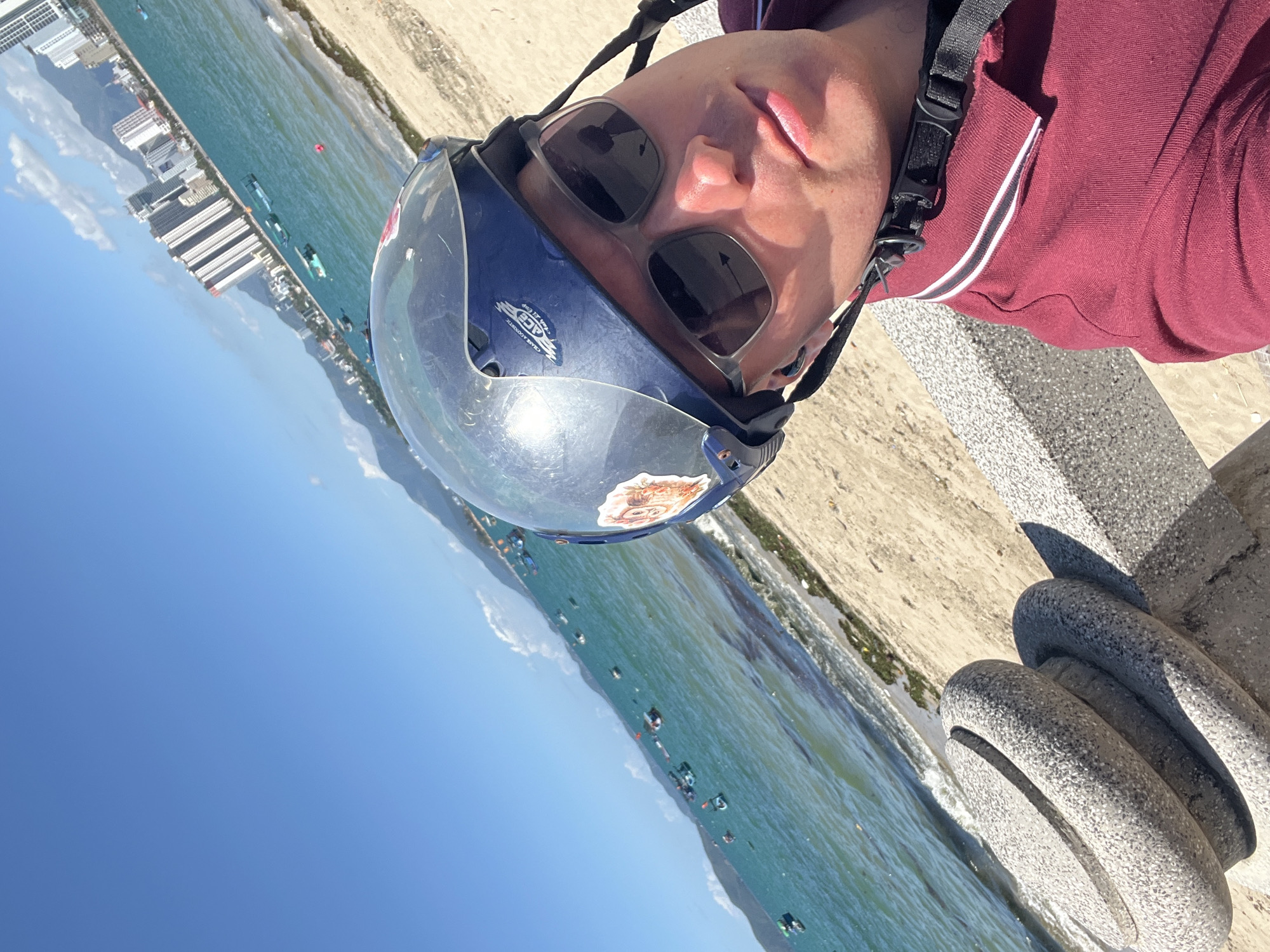 A person wearing sunglasses and a blue helmet with a clear visor is positioned in the foreground. They are wearing a maroon polo shirt with white stripes on the collar. The background shows a view of the ocean with several small boats. The coastline features a beach and buildings, suggesting an urban area. The location appears to be near the water, with a concrete structure visible near the bottom right. The image is taken in Bac Nha Trang Ward, Vietnam.