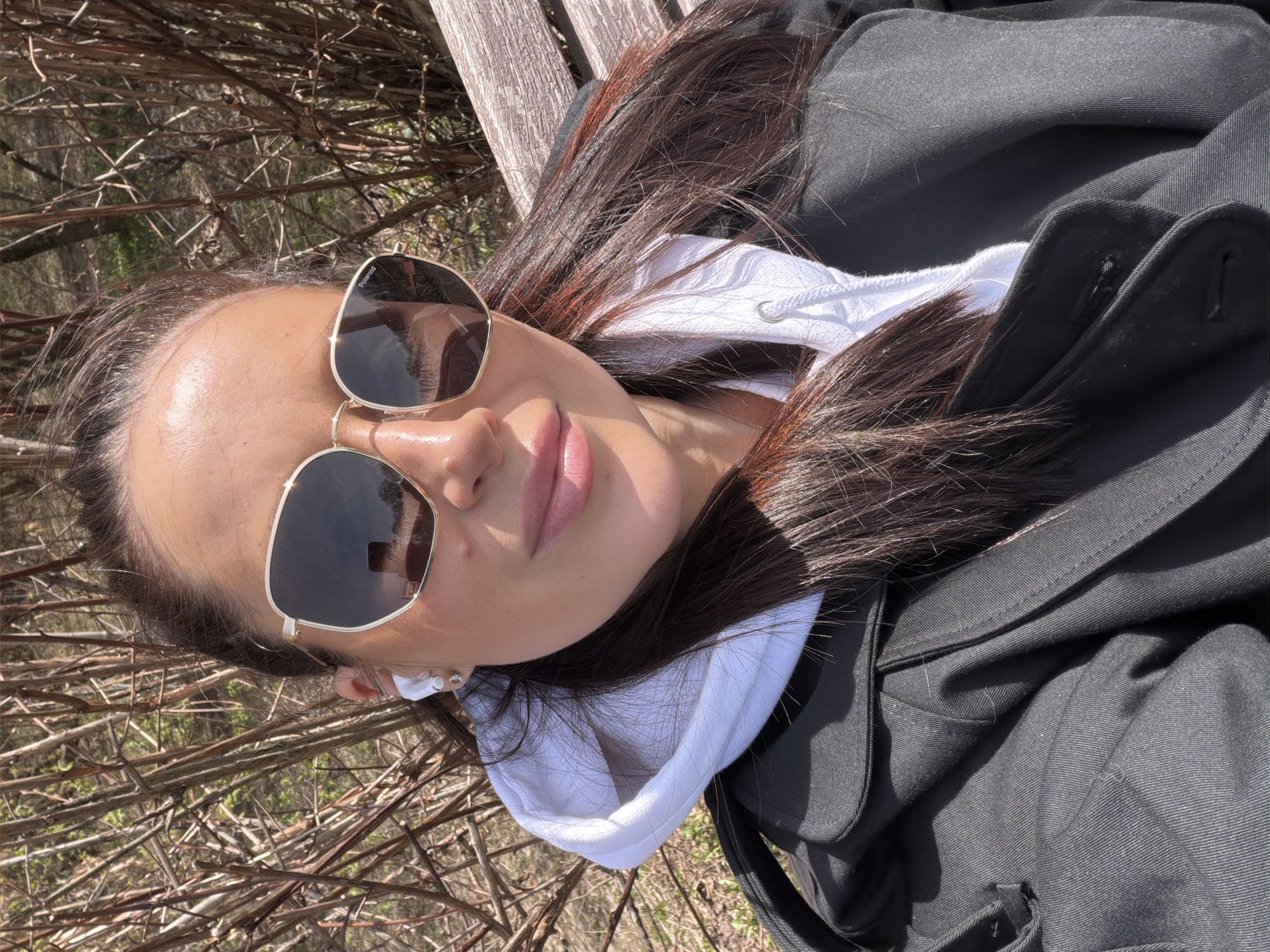 A medium shot depicts a woman wearing sunglasses and a white hoodie under a dark jacket, lying on a wooden surface that appears to be a bench. The background consists of bare branches and vegetation, suggesting an outdoor location. The woman is wearing an earpiece. There is no information available about the location being in Khimki, Russia in the image.