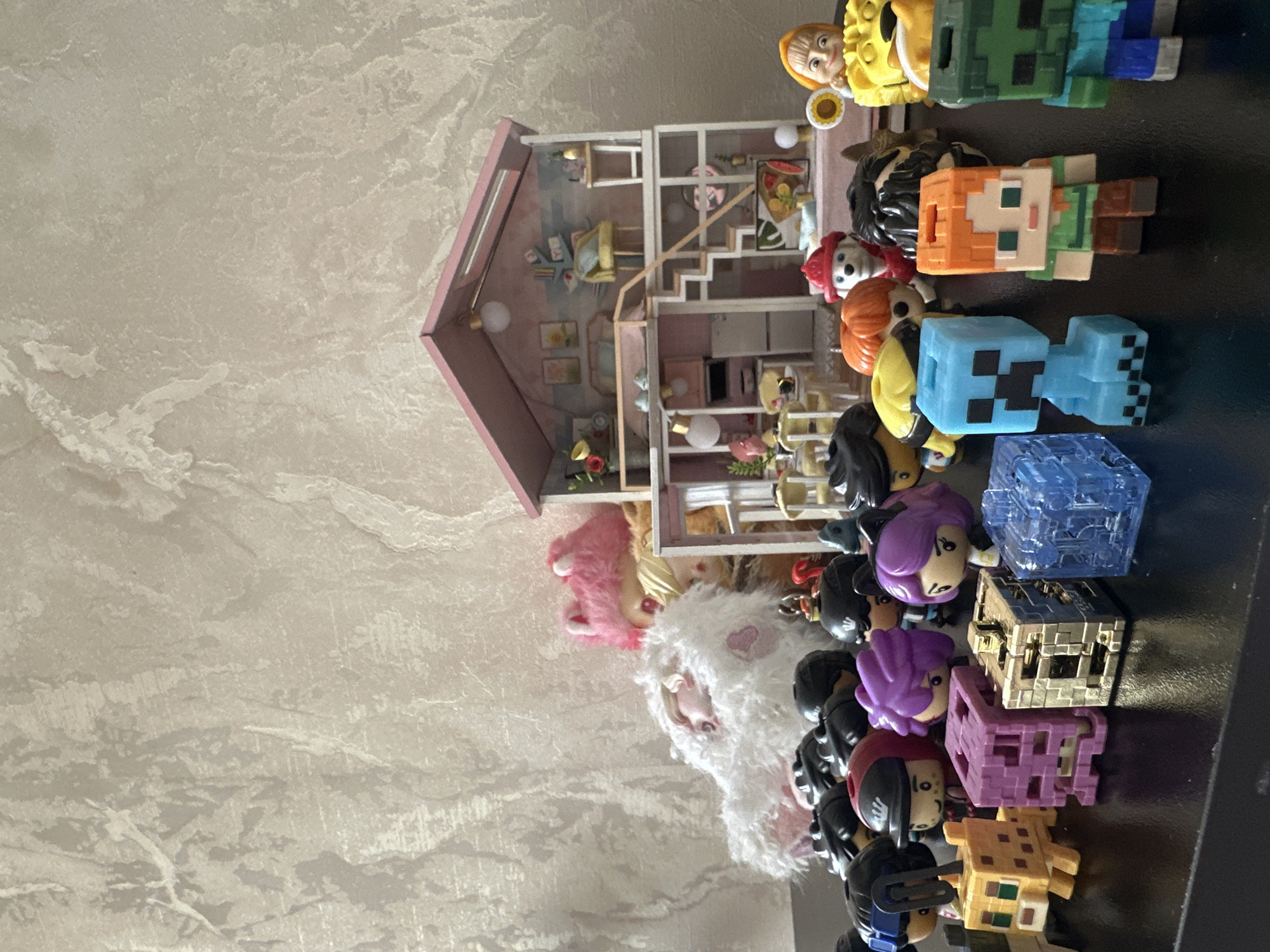 The image shows a collection of toys arranged on a surface with a textured, stone-like appearance, possibly a countertop or table. The toys include a pink dollhouse, several figurines that appear to be from the Minecraft video game, and other smaller dolls or figures. The scene is cluttered, with the toys arranged closely together. There's no evidence of Yerevan, Armenia, in the image.