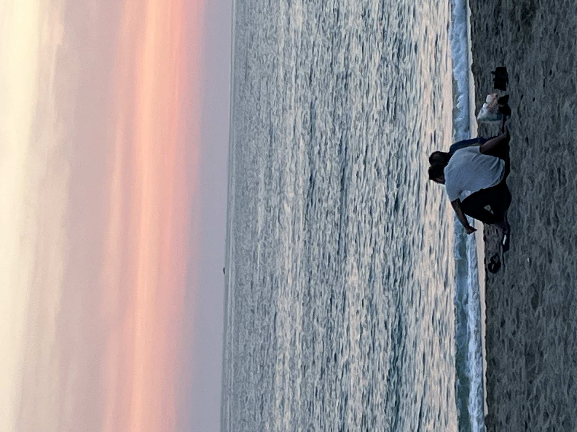The image depicts a beach scene, likely at sunset, in Alanya, Turkey. A couple is seen on the sand near the water's edge. One person is wearing a white shirt and dark pants. They appear to be interacting with the sand or water. Various items, including a camera, are scattered nearby on the beach. The sea is calm, and the sky exhibits a gradient of colors, from light orange to pale yellow.