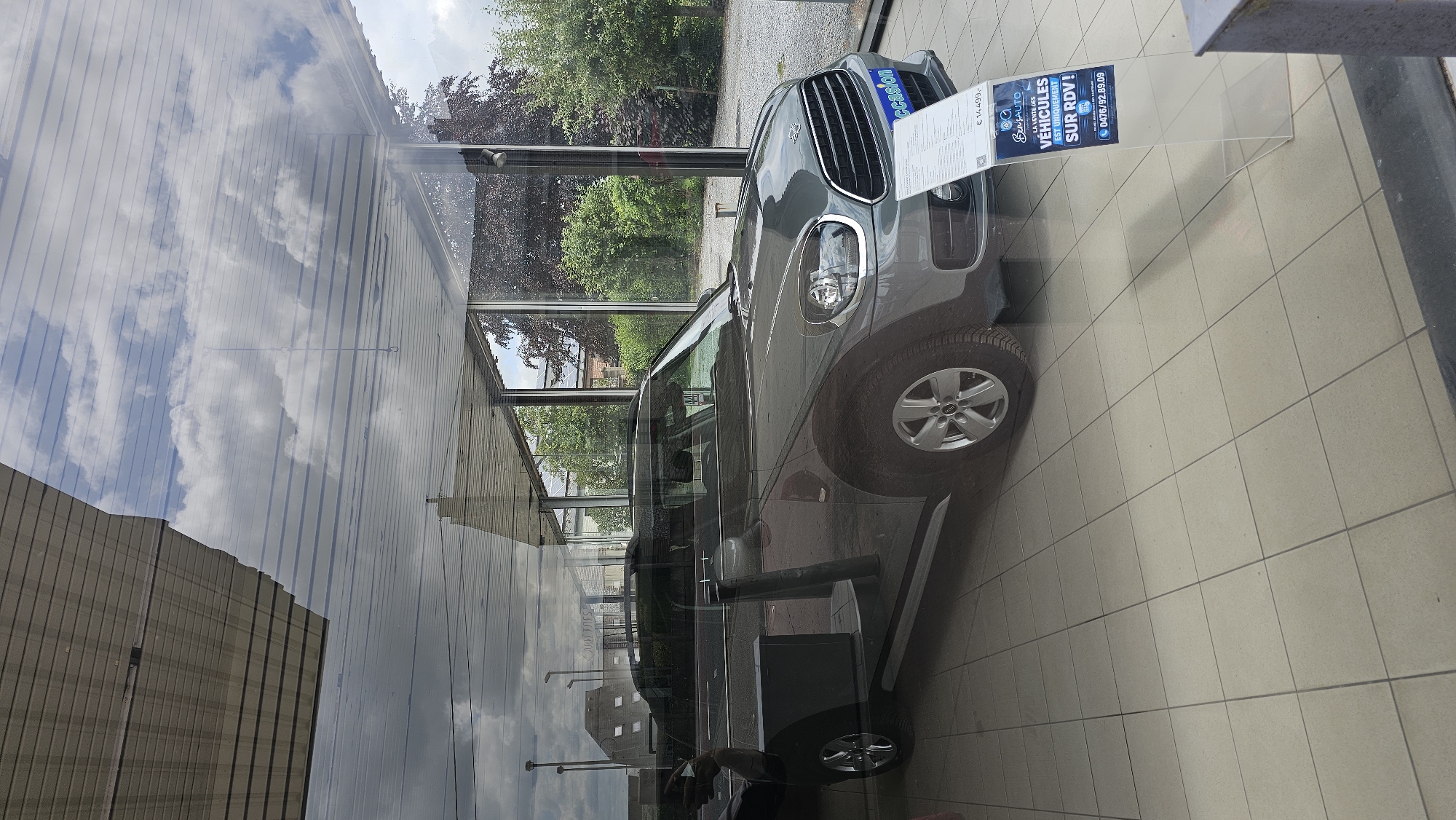 An exterior shot, taken through a window, shows a grey Mini Countryman on display in what appears to be a car dealership in Boussu-lez-Walcourt, Belgium. The car is positioned at an angle, with its driver's side door open. The car's interior is visible, as are its headlights and grille. A sign in front of the car displays the price "€ 14.499,-" and contact information. The car is parked on a tiled floor. Reflections on the window reveal a cloudy sky and the dealership's roof structure. Outside the dealership, trees and buildings can be seen.