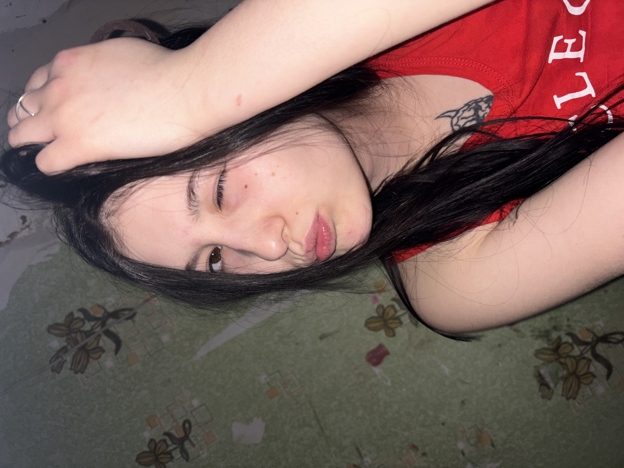 A close-up, indoor portrait depicts a young female individual with long, dark hair and fair skin. Her right eye is closed in a wink, while her left eye is open and directed towards the viewer. Her lips are puckered into a "kiss" pose. Her left hand is raised, with fingers positioned in her hair, and a plain silver-colored band ring is visible on her left ring finger. A faint reddish mark is present on her left forearm. A dark, stylized tattoo, appearing to be a horned figure or animal head, is visible on her upper left chest/shoulder, partially obscured by her hair and a red tank top. The tank top displays white lettering, with "LE" visible on the lower right portion of the garment. The background consists of a wall covered in light green wallpaper featuring a repeating pattern of stylized light brown floral and geometric designs. The wallpaper exhibits signs of wear, including peeling and discoloration. The scene is purportedly located in the city of Foki, Russia.