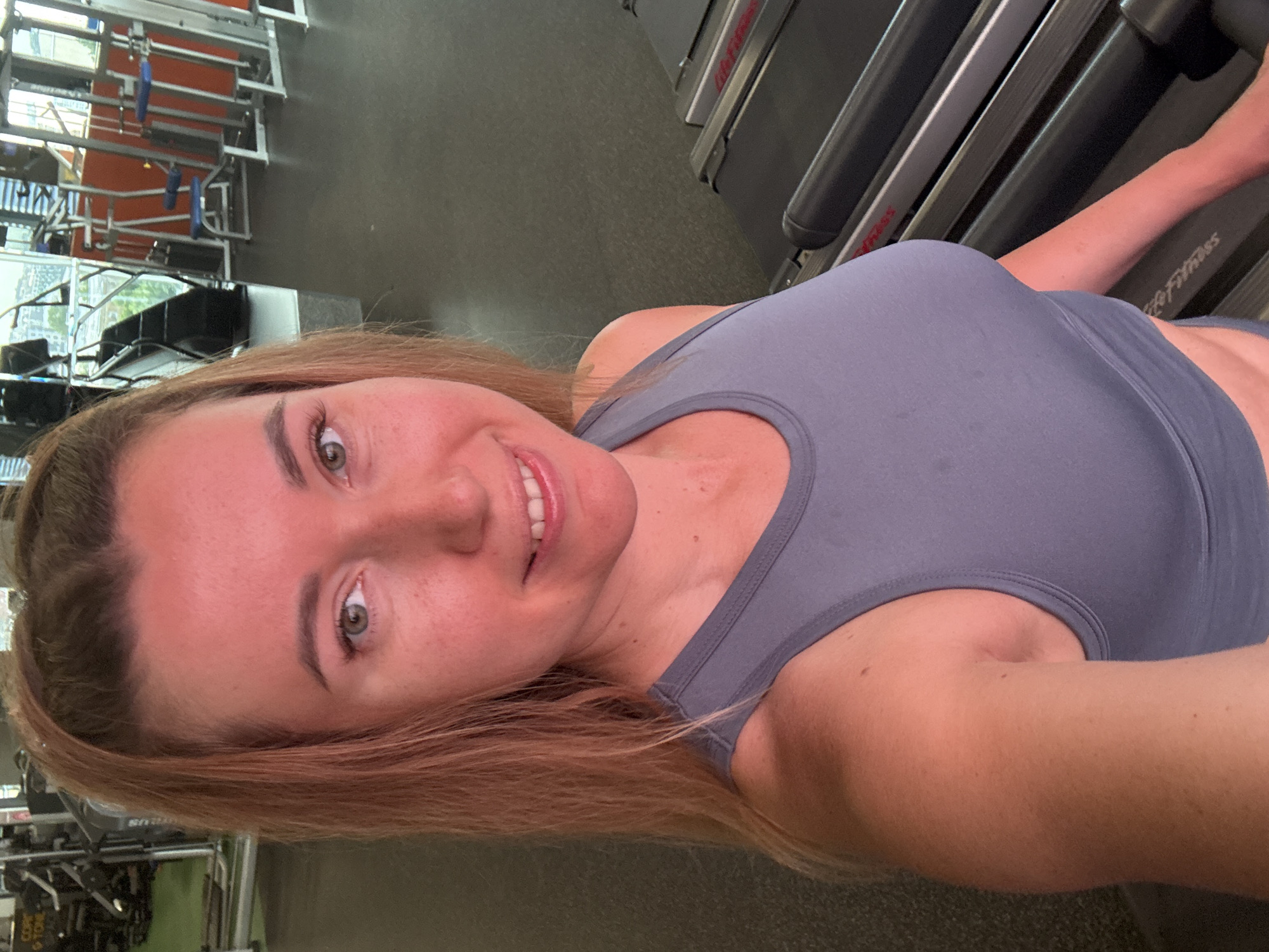 A woman is taking a selfie in a gym. She is wearing a grey sports bra and has her hair pulled back. She is smiling at the camera, and her face is flushed, likely from exercise. Behind her, treadmills are visible. The gym appears to be well-lit and equipped with various exercise machines. The overall atmosphere is active and fitness-oriented.