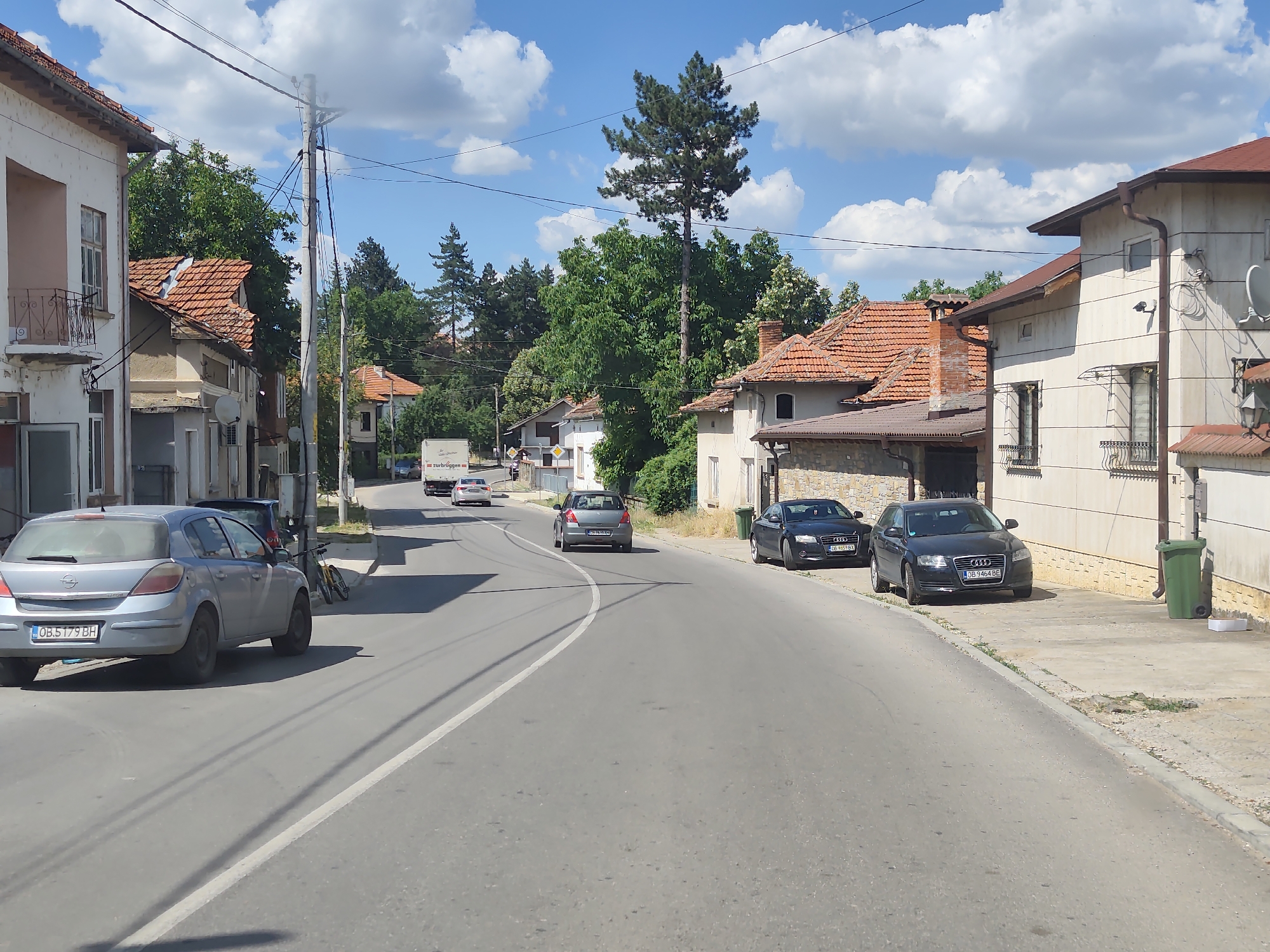 This media file captures a daytime scene in Dermantsi, Bulgaria, on a sunny day with scattered clouds. The image depicts a street lined with buildings and trees. On the left side of the road, a light blue Opel Astra hatchback is parked. Further down the street, several other cars are visible, including a dark grey sedan and two black Audi sedans parked on the right side. A large green trash bin is also present near the Audi sedans. In the background, a white truck with the text "ZURBRIGGEN" is partially visible. The architecture of the buildings suggests a European setting, with tiled roofs and balconies. The road itself is paved and curves gently to the right. Overhead, power lines crisscross the sky. The overall impression is of a quiet, residential street on a clear day.