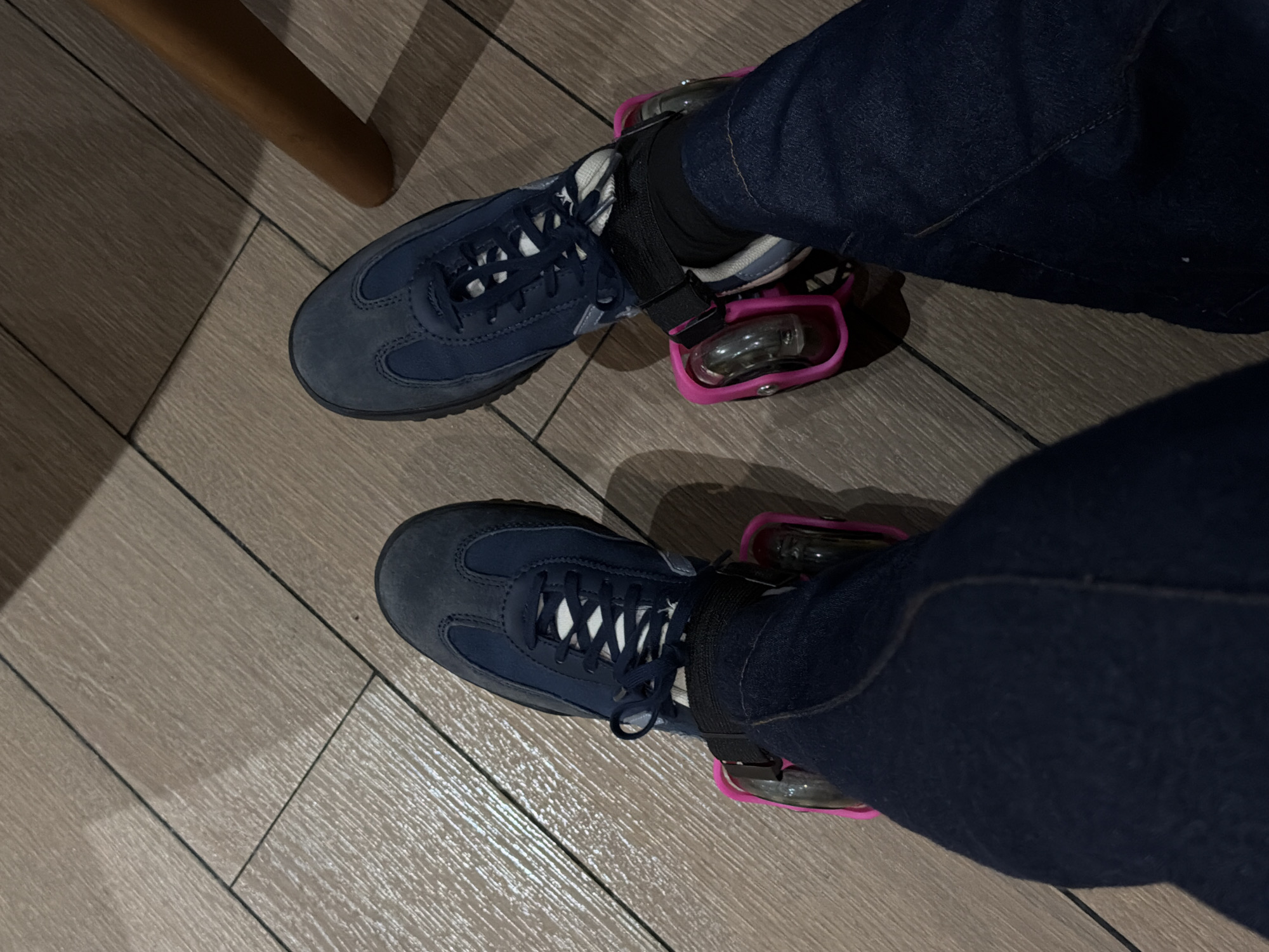 The image shows a person's legs and feet, wearing blue sneakers with pink roller skate attachments. The person is standing on a wooden floor. The scene is dimly lit, suggesting an indoor environment, possibly a home or a public space. The roller skate attachments have two wheels each. The person is wearing dark blue jeans. The image is taken from a high angle, looking down at the person's feet.