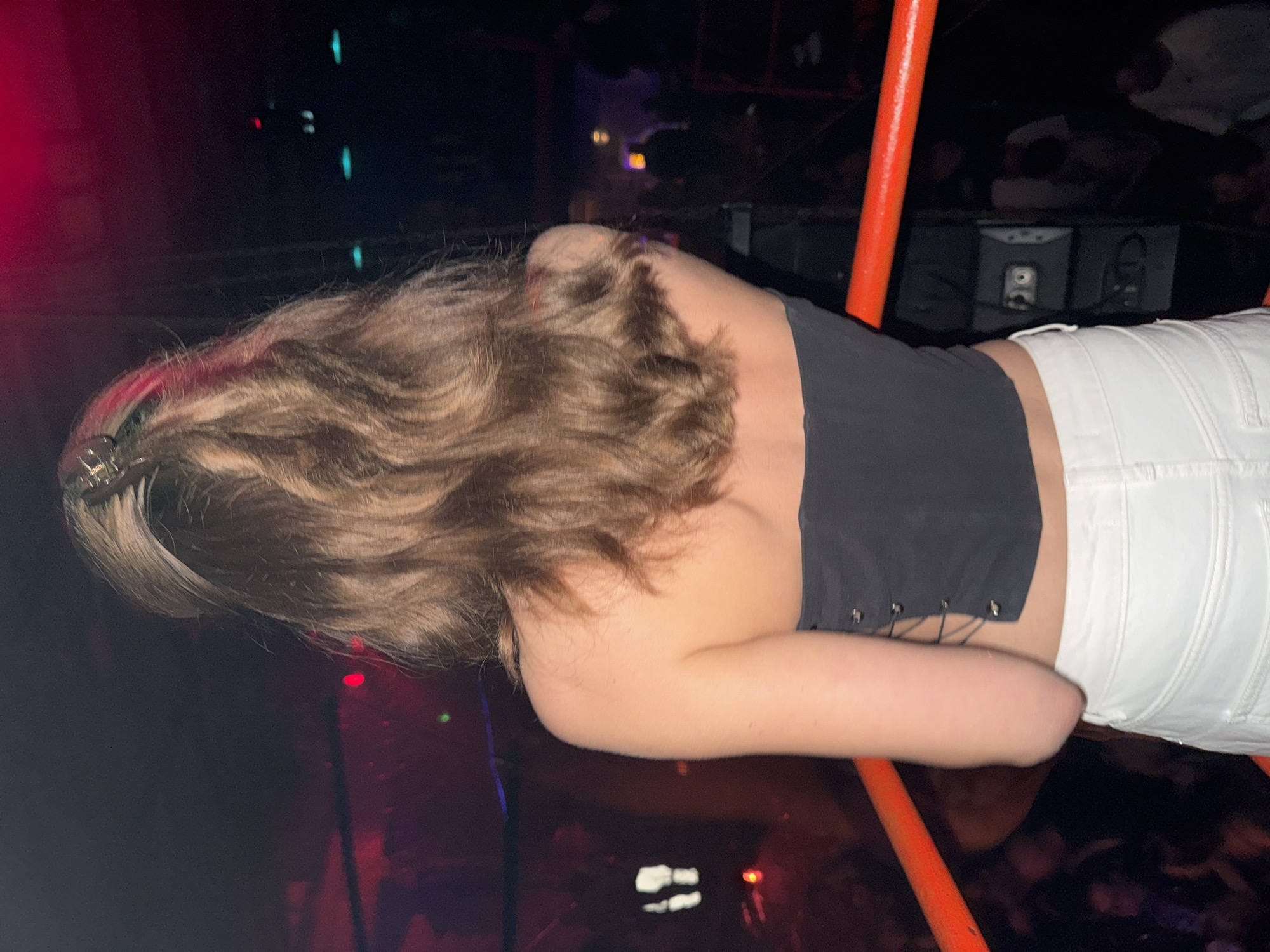 A woman with brown hair and a hair clip, wearing a black top and white pants, leans over a dark surface with an orange pole beside her. The background is dark, with hints of lights and a crowd. The location is indiscernible from the provided image.