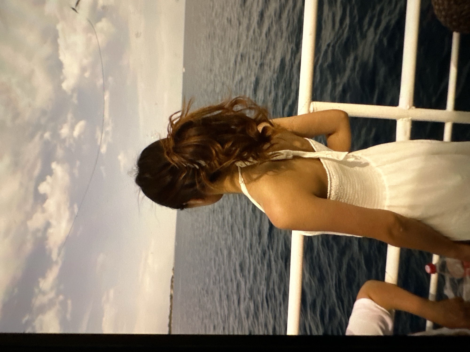 A woman with auburn hair pulled into a high ponytail stands with her back to the camera, wearing a white sundress. She is on a boat, leaning against a white railing. The sea is visible behind her, with a distant shoreline. The sky is cloudy and overcast. The scene takes place outdoors, on the water. There is a bottle of water on the deck of the boat. The specific location in the city of Кызыл-Булак, Kyrgyzstan, cannot be determined from the image.