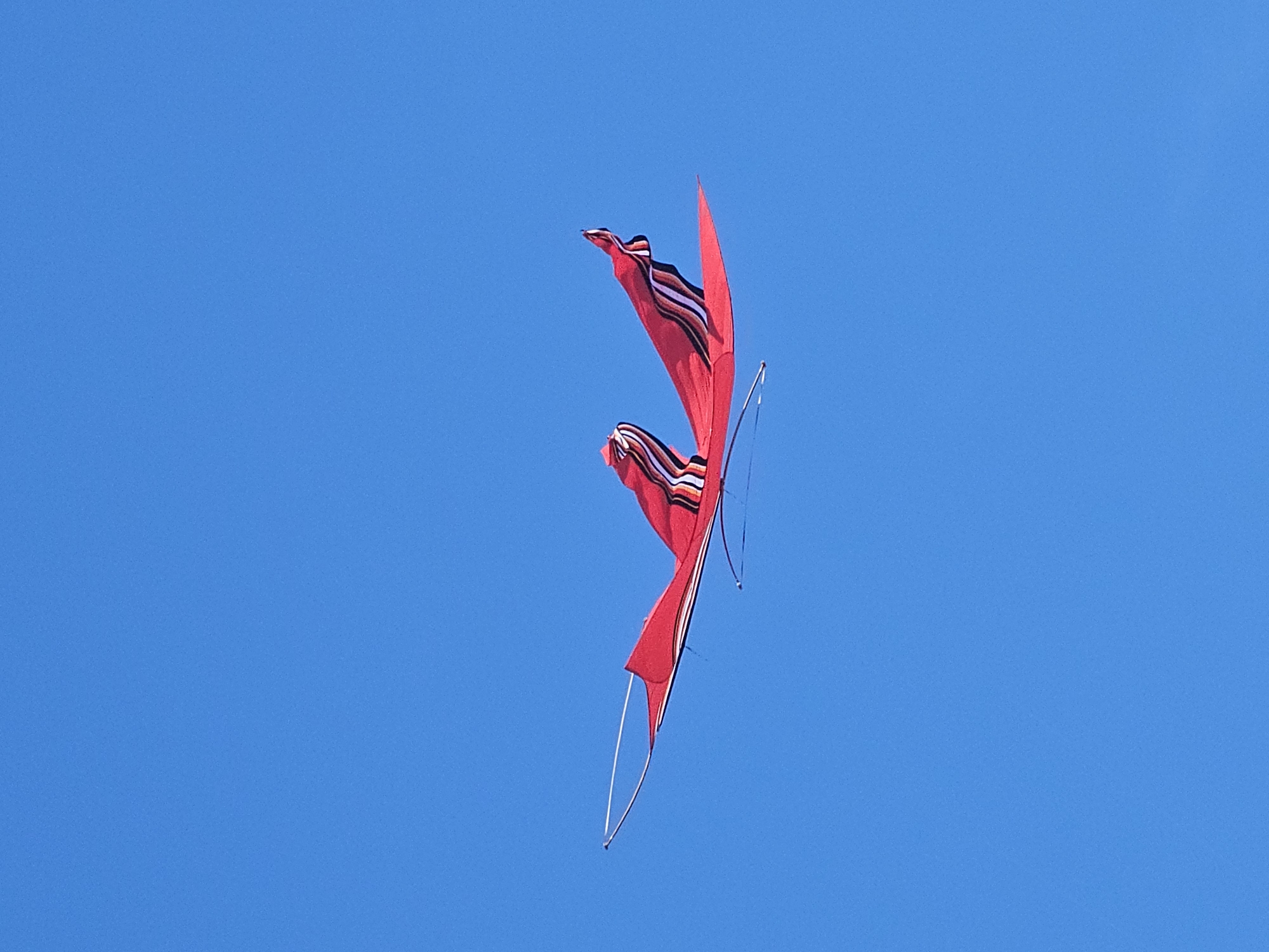 A red, Balinese kite is soaring against a cloudless, blue sky, presumably in Denpasar, Indonesia. The kite has a distinctive design with red fabric and black and white striped accents. Thin white rods frame the kite's edges.