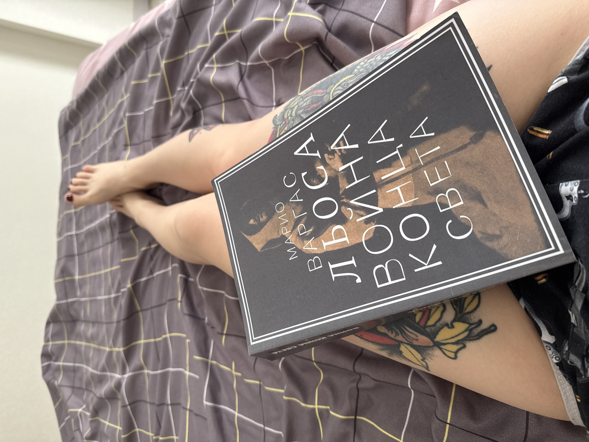 A person with tattoos on their legs is lying on a bed with a gray and yellow checkered pattern comforter. A book with text in a Cyrillic script is placed on top of the person's legs. The edges of the book have a thin white border. The person is wearing dark-colored pajamas with a pattern. The background is blurred. The location is unknown.