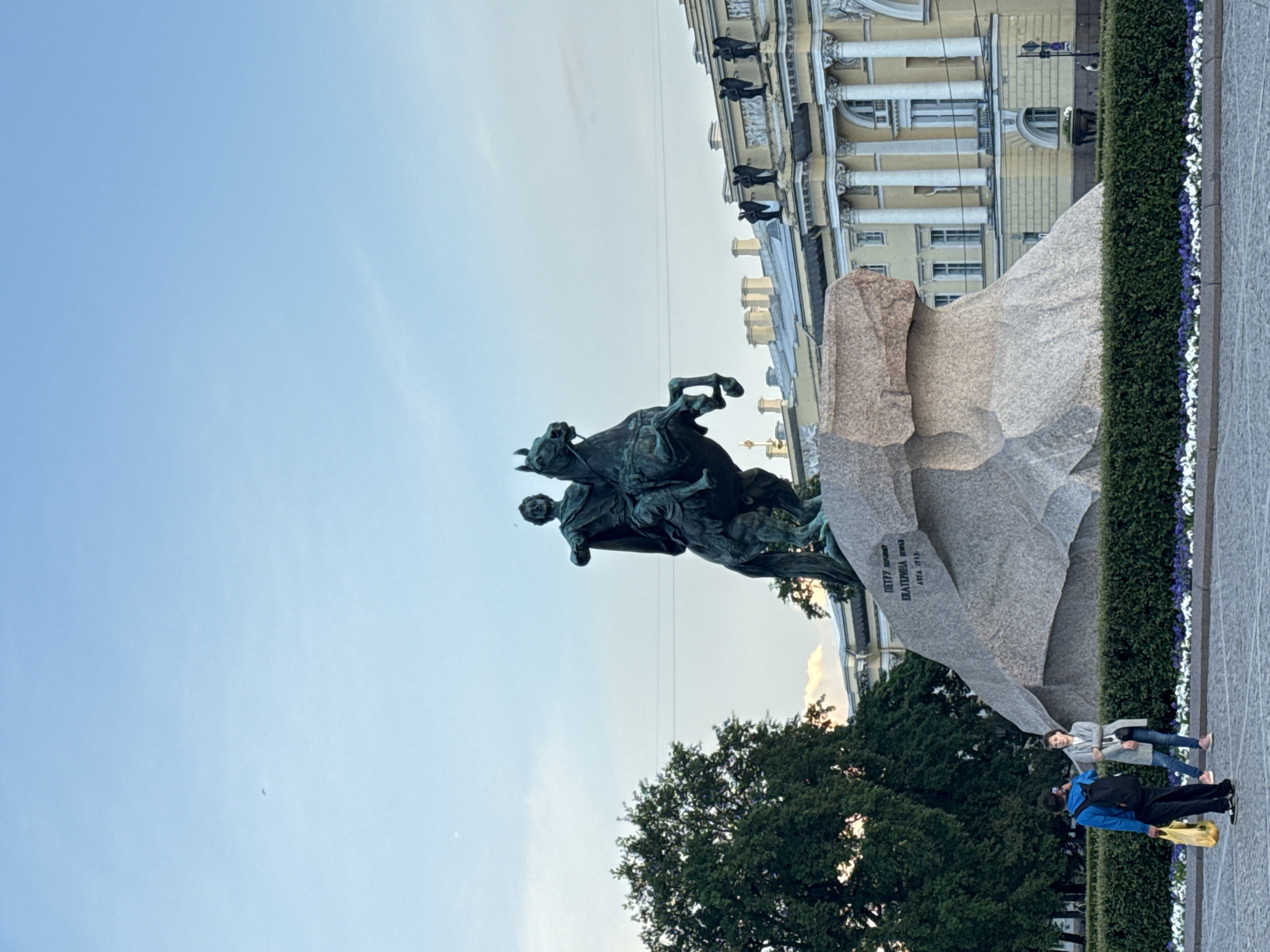 The image shows the Bronze Horseman, a monument to Peter the Great, located on Senate Square in Saint Petersburg, Russia. The equestrian statue is depicted in mid-gallop, with the horse rearing up on its hind legs. The base of the statue is a massive stone, known as the Thunder Stone, which is inscribed with text. In the foreground, two individuals are visible. One person, wearing a blue jacket and black pants, is standing with their back to the camera, holding a yellow bag. The other person, dressed in a grey coat and blue jeans, is facing towards the monument and holding a handbag. Behind the monument, a large, ornate yellow building with columns and statues on its roof is visible, likely part of the administrative buildings surrounding Senate Square. Lush green trees are present on the left side of the image, and a manicured hedge with flowers lines the front of the scene. The sky above is a clear blue with some faint clouds.