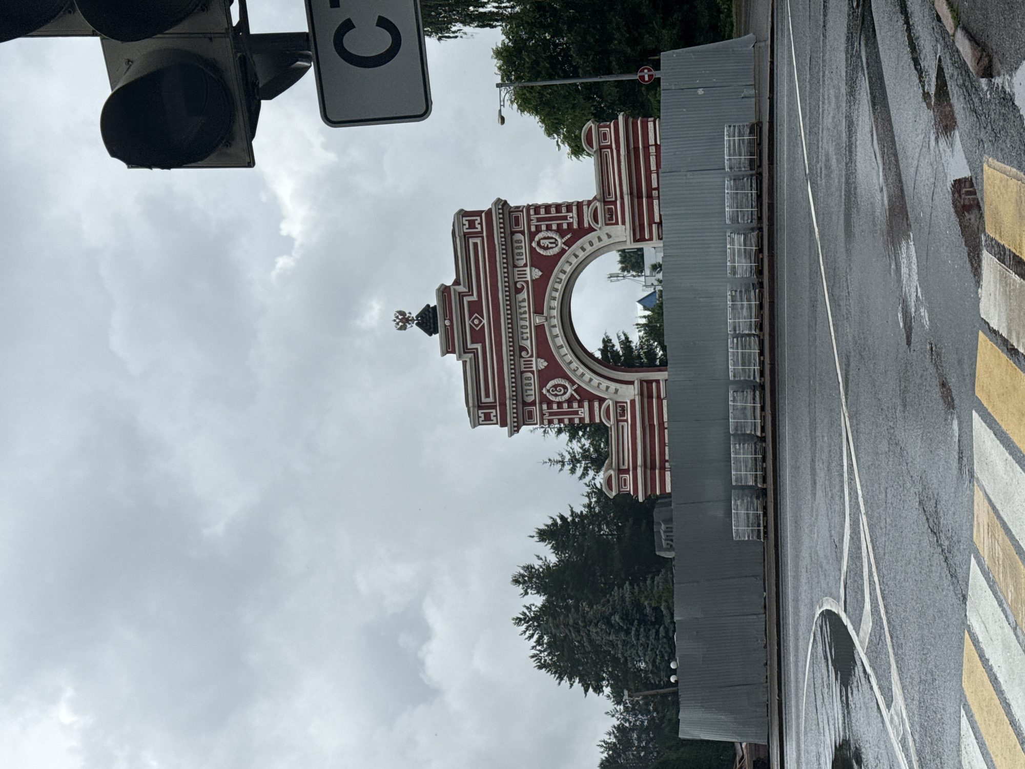 The image captures a red and white archway in Kazan, Russia, under a cloudy sky. The archway is decorated with historical inscriptions and a double-headed eagle emblem at its apex. In front of the arch, a temporary gray metal fence encloses a construction area, with stacks of packaged paving stones visible on pallets behind the fence. A traffic light and a street sign with a black "C" on a white background are present to the right of the arch. The ground in the foreground is wet, indicating recent rain, with a pedestrian crossing marked by yellow and white stripes. Trees and buildings are visible in the background.