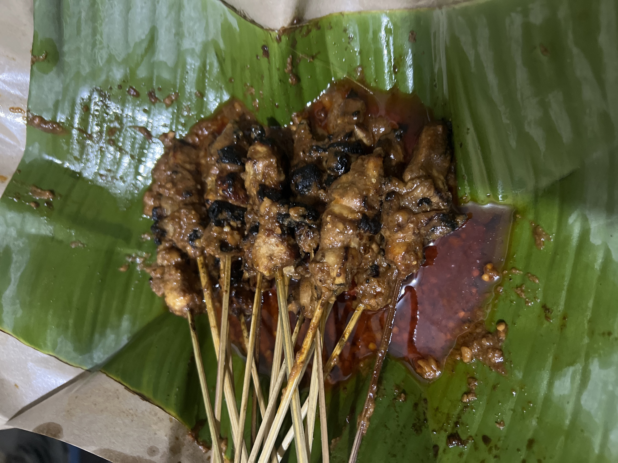 A close-up, overhead shot shows a serving of grilled meat skewers, likely satay, presented on a large, vibrant green banana leaf. The skewers, made of thin wooden sticks, are laden with chunks of seasoned meat that appear to be grilled to a slightly charred finish. A rich, reddish-brown sauce, possibly peanut-based, pools around the skewers and also coats the meat. Some of the sauce has dripped onto the banana leaf, creating glossy streaks. The banana leaf is slightly wilted and has a waxy sheen, suggesting it is fresh. The background consists of crumpled brown paper, hinting at packaging or serving material. The image appears to have been taken indoors under artificial lighting, judging by the way the light reflects off the glossy sauce and the banana leaf. The setting, based on the food presentation, is likely a street food stall or casual eatery in Klapanunggal, Indonesia, a region known for its diverse culinary offerings. There are no people or animals visible in the frame. The focus is entirely on the appetizing presentation of the food.
