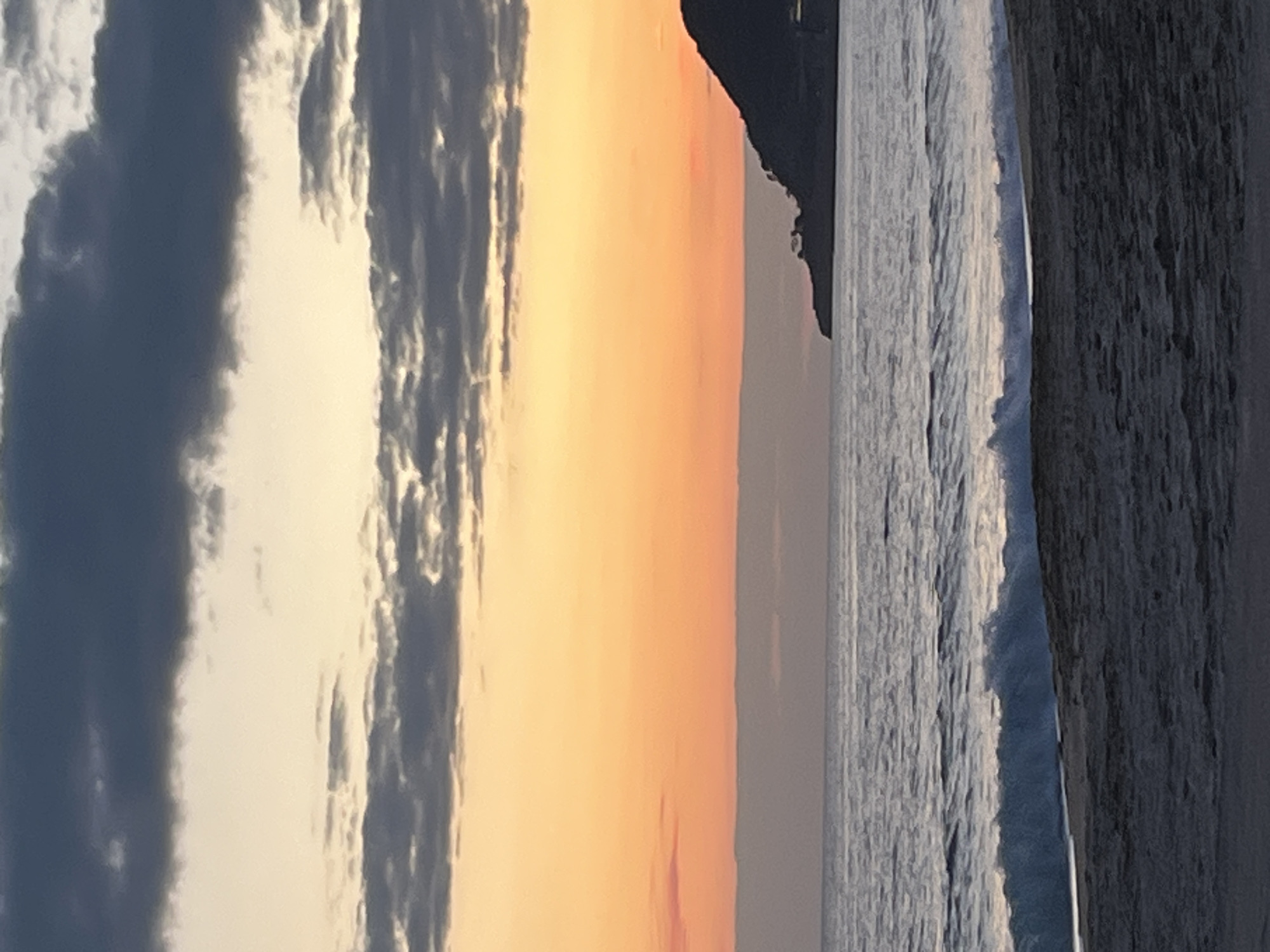 Here is a description of the image.
The image presents a view of the shoreline during sunset in Alanya, Turkey. The sky features a band of orange light, indicative of the setting sun, flanked by dark, partially cloud-covered areas. The sea is visible, showing wave activity. A dark landmass is present between the sunset and the sea. A pier or the edge of a structure is on the edge. No people are visible in the image.