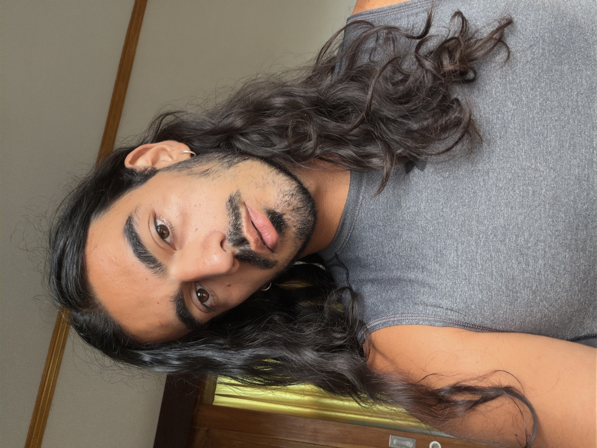 The image depicts a person with long, wavy dark hair wearing a gray tank top. The individual has facial hair and an earring in their left ear. The background includes a wall, a wooden trim, and what appears to be a wooden door or frame. The location is suspected to be in North Kuta, Indonesia, but there are no geographical markers to positively confirm this. The person is rotated in the image.